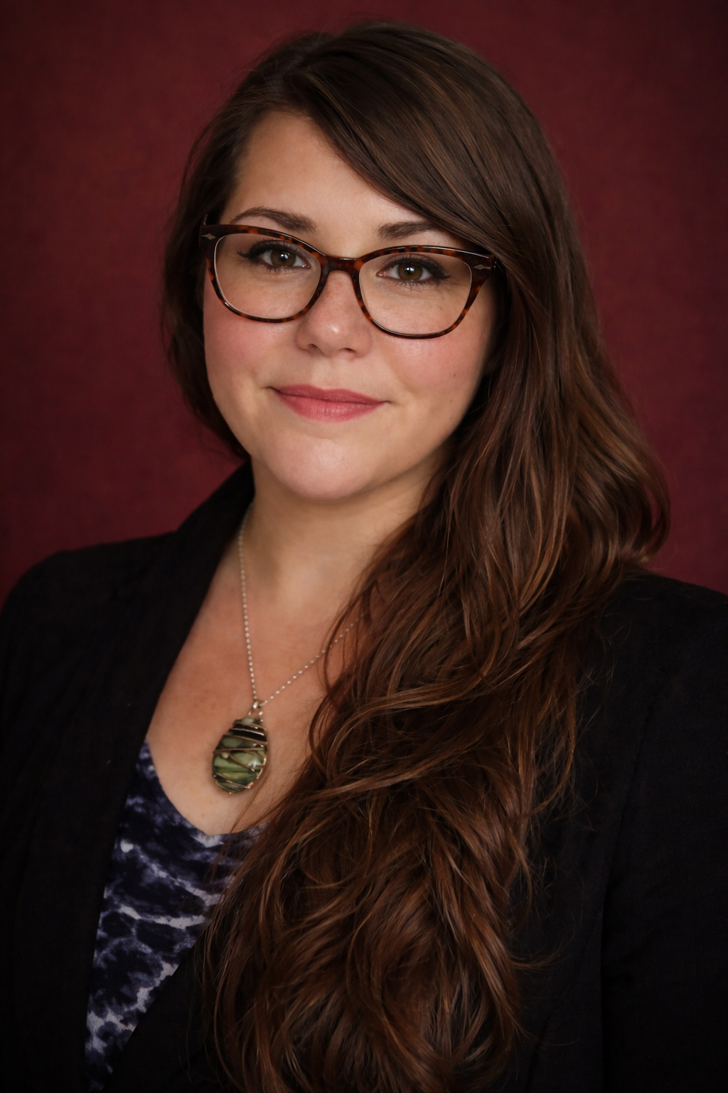 Portrait of a woman with long, wavy brown hair wearing glasses, a black blazer, a patterned top, and a necklace with a green and black pendant, against a dark red background.
