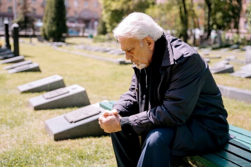 Hombre mayor triste con la cabeza gacha sentado en un cementerio y necesitando coaching