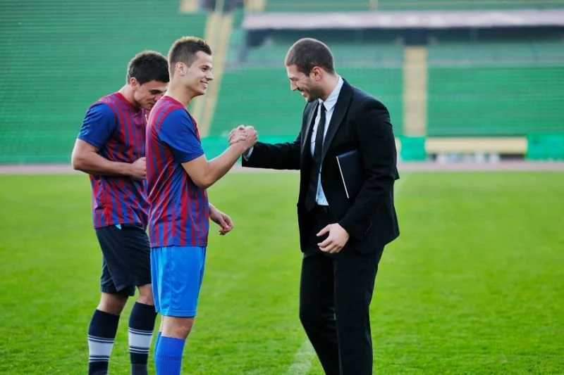 Coach deportivo con saco y corbata saludando a jugadores de fútbol luego de una sesión