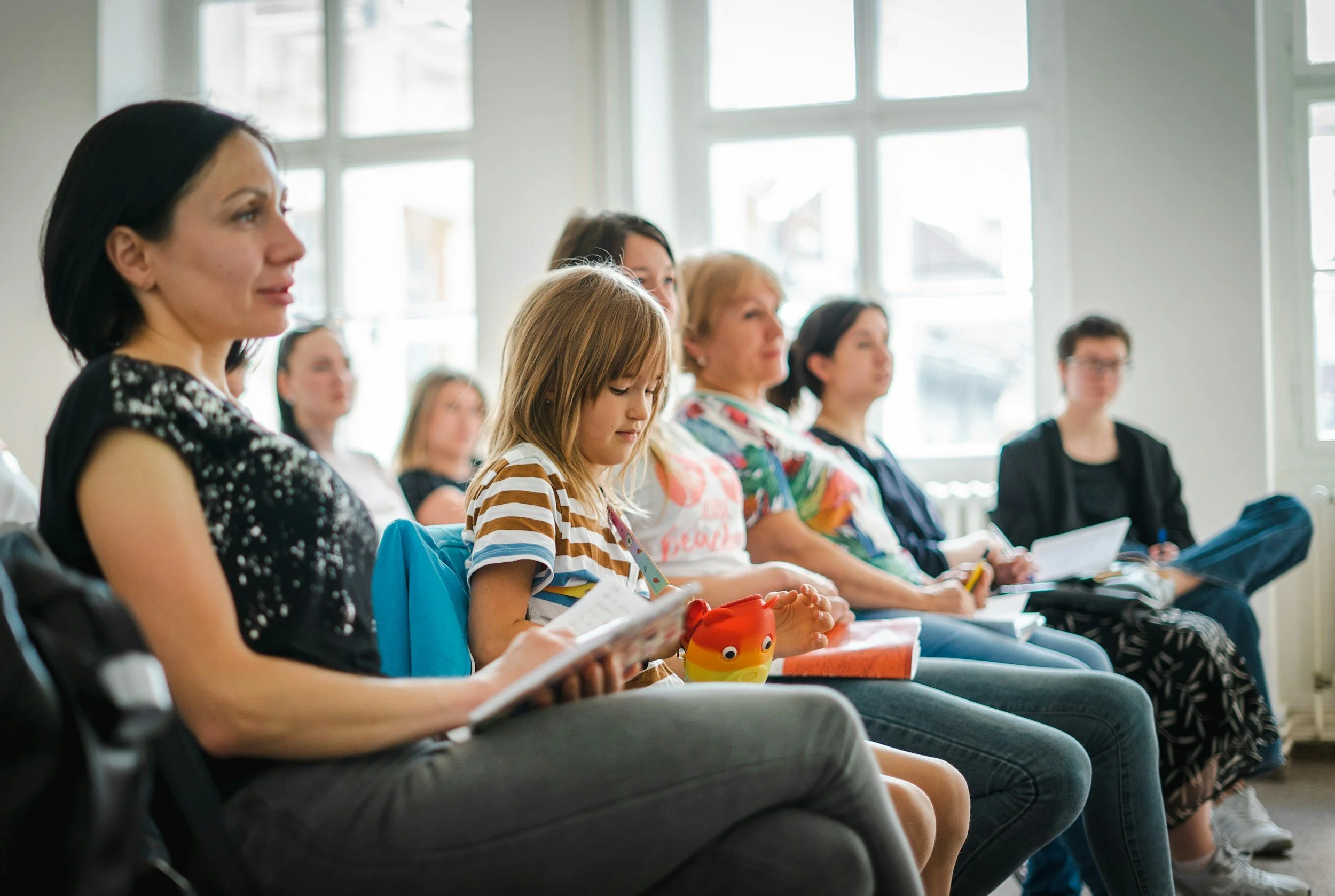A group of women and children seated indoors, listening attentively during an event or class, with large windows in the background.