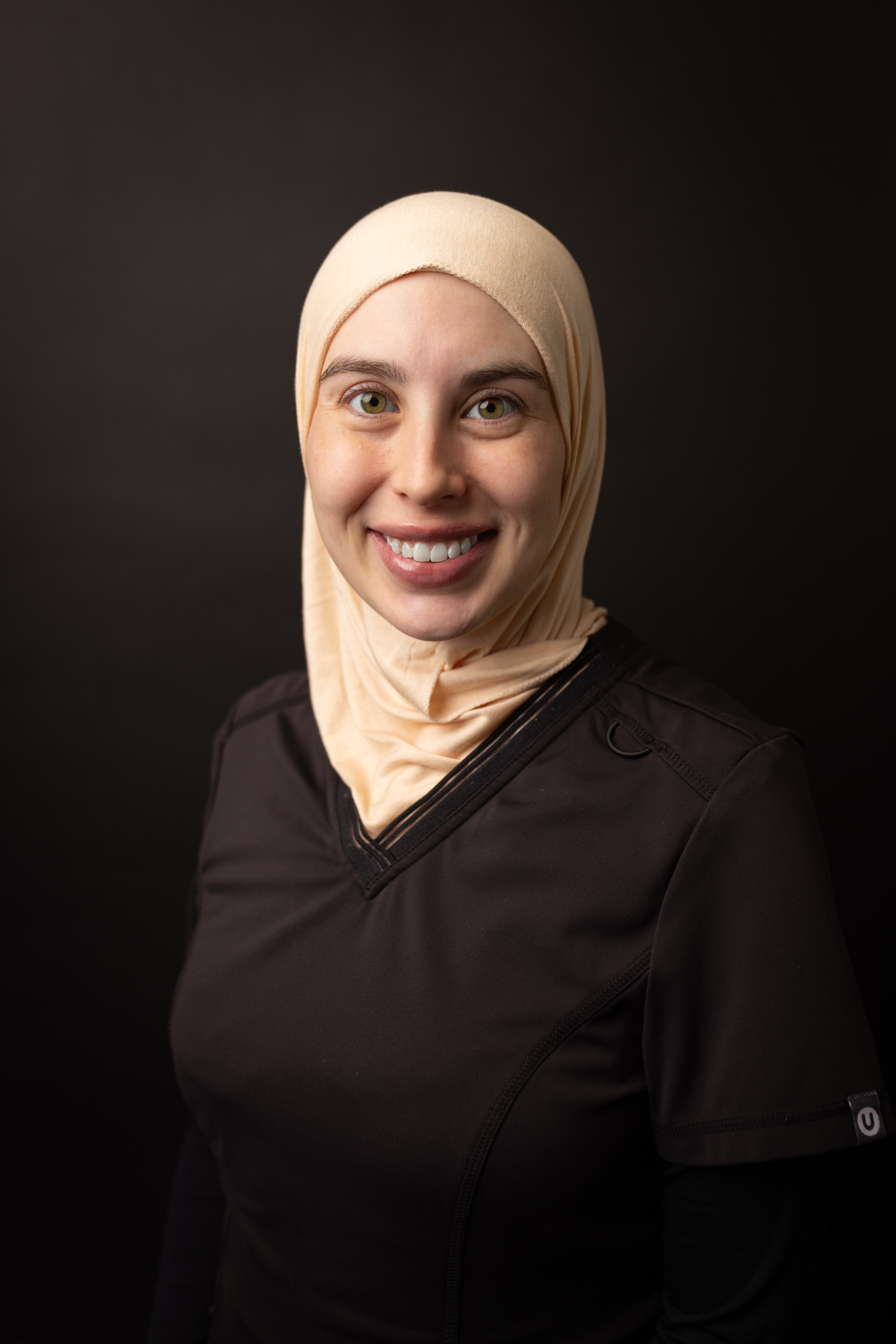 Portrait of a smiling woman wearing a beige hijab and a black jacket against a dark background.