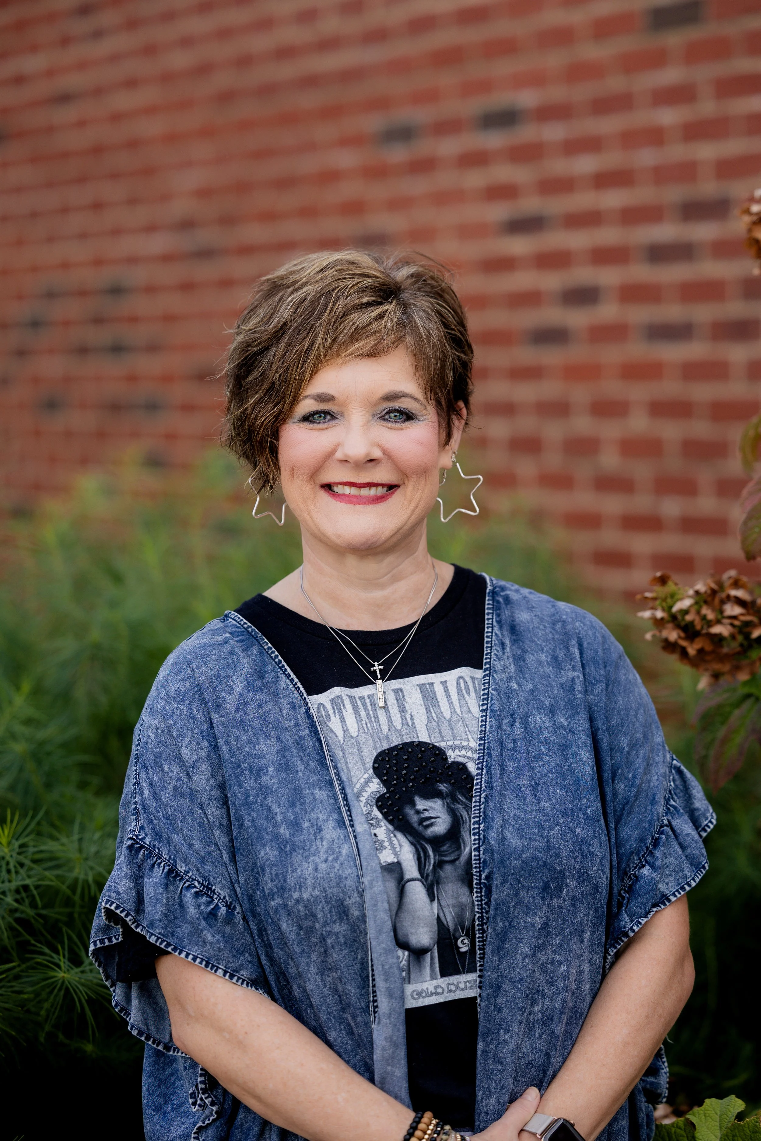 Smiling woman with short curly brown hair wearing a black top with studded details around the neckline and sleeves, light blue jeans, and jewelry standing outdoors in a wooded area.