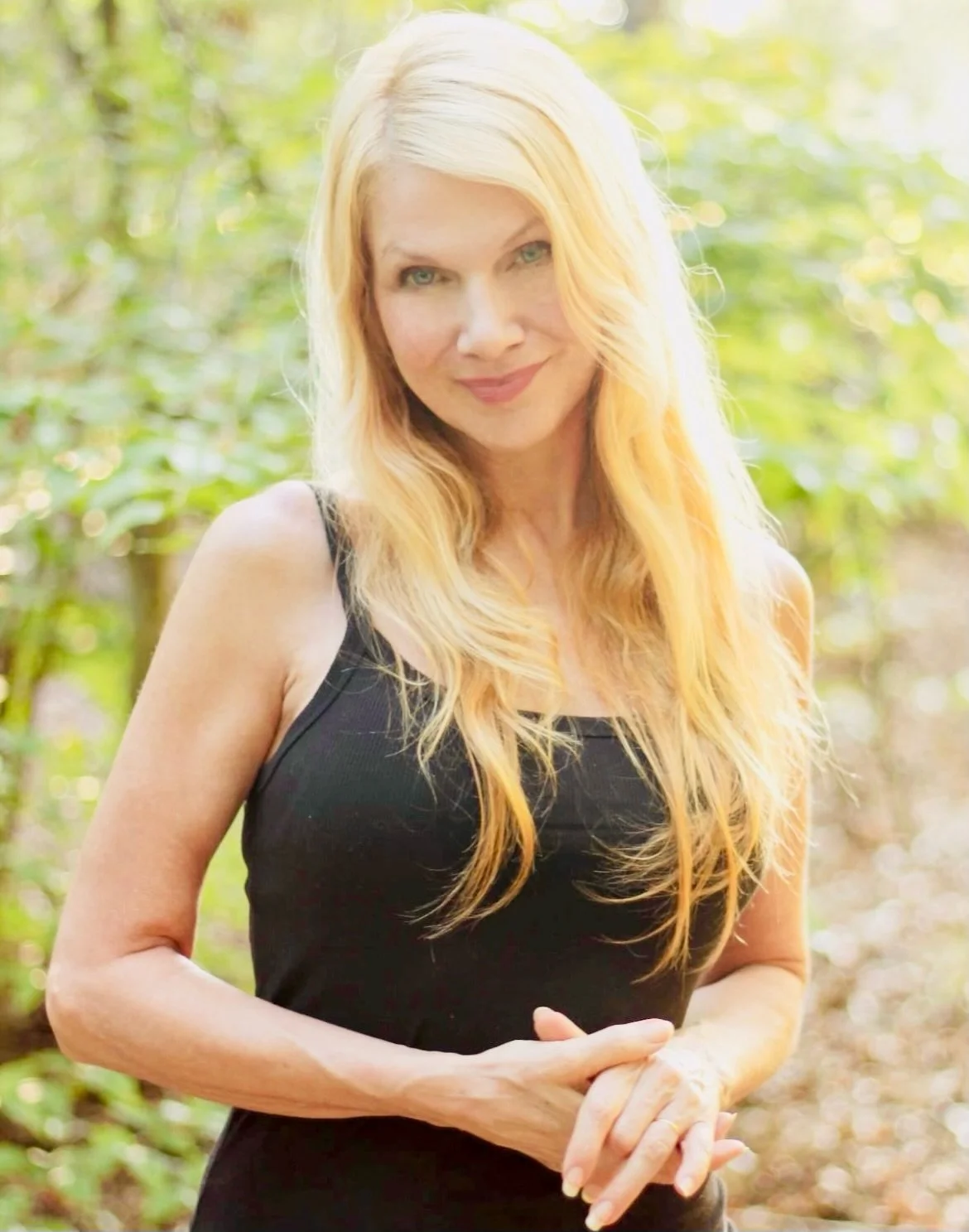 A woman with long blonde hair smiling and holding sunflowers in a field of sunflowers during daytime.