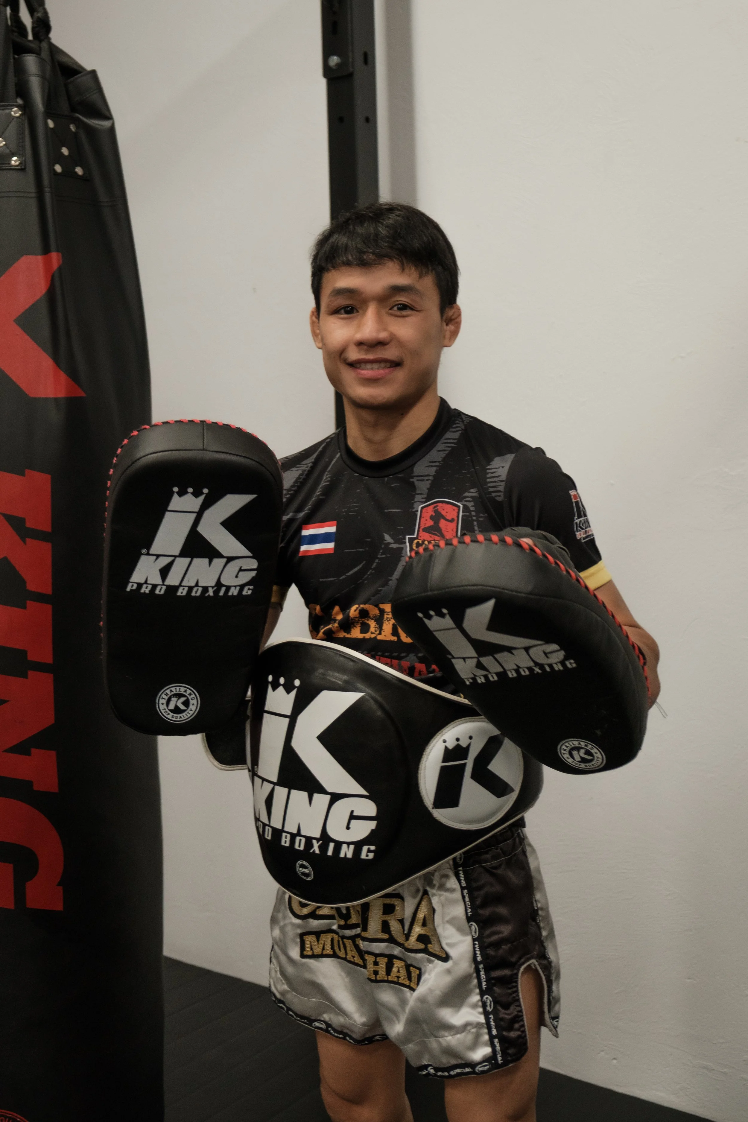 Young male boxer in black and gold Muay Thai shorts and black gloves, standing inside a boxing gym, smiling, with a white wall and punching bag in the background.
