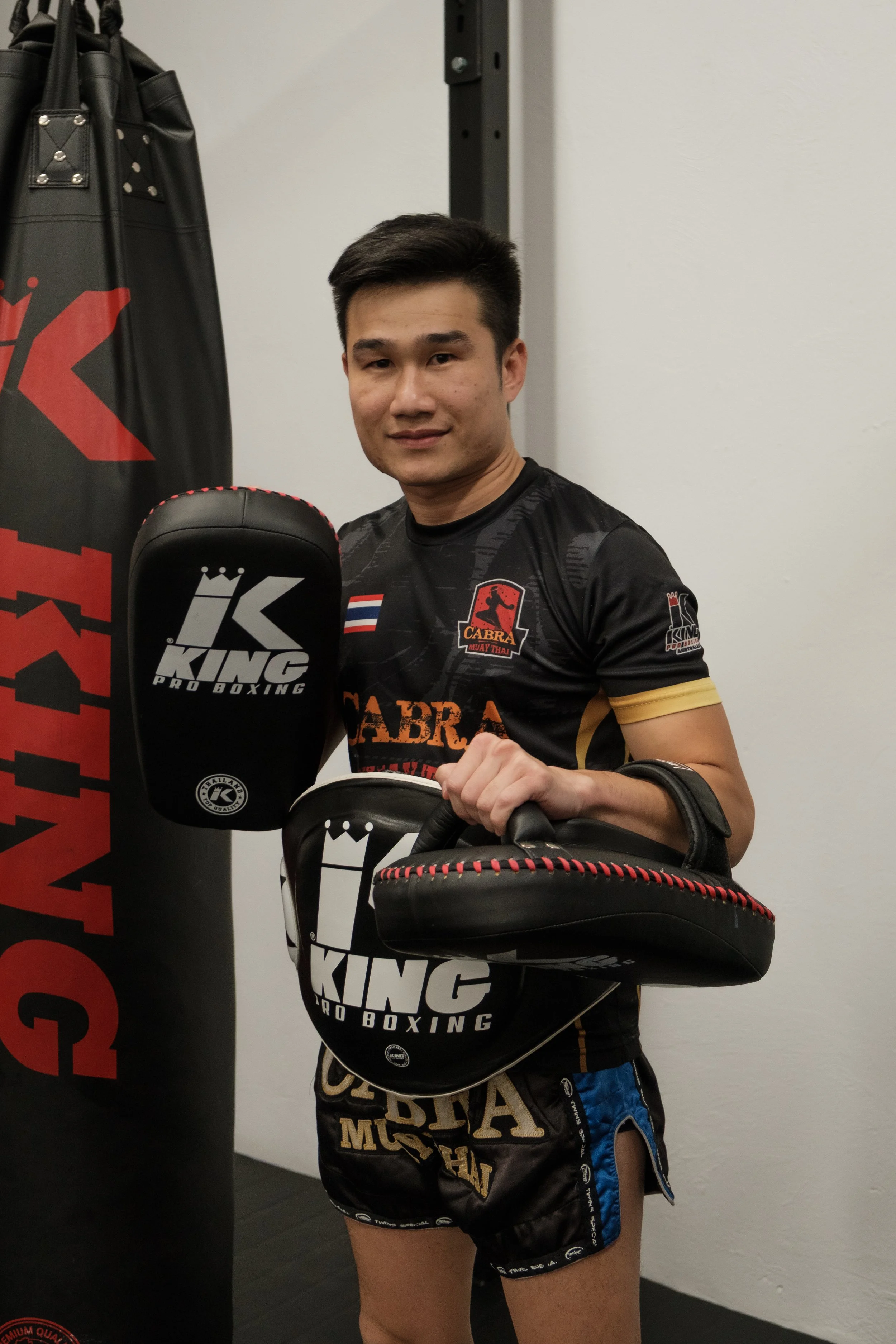 A male Muay Thai fighter holding focus mitts and standing next to a boxing training bag, wearing a black and gold uniform with logos.