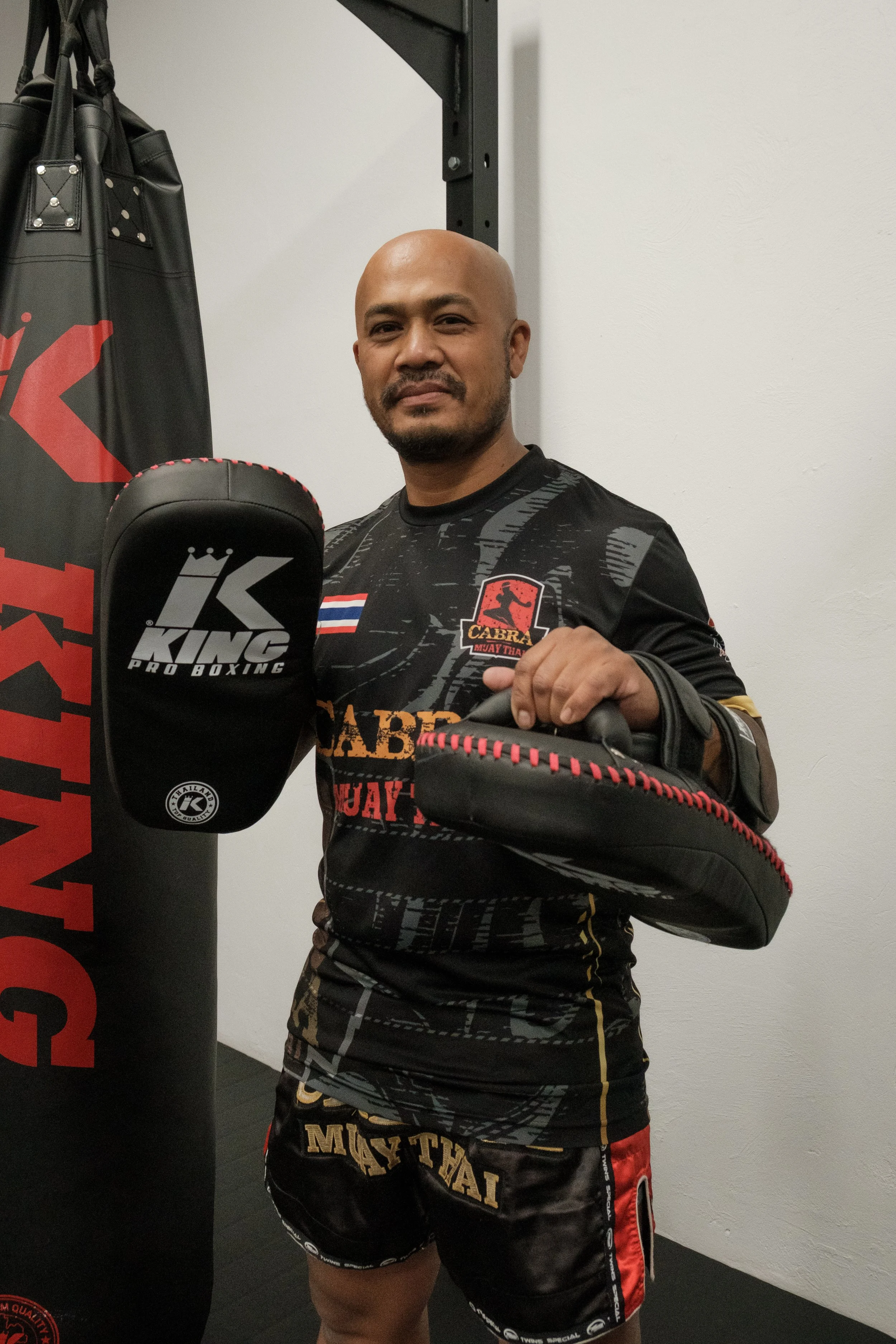 A man standing in a martial arts gym poses with his fists raised, wearing hand wraps and martial arts attire, in front of a punching bag.