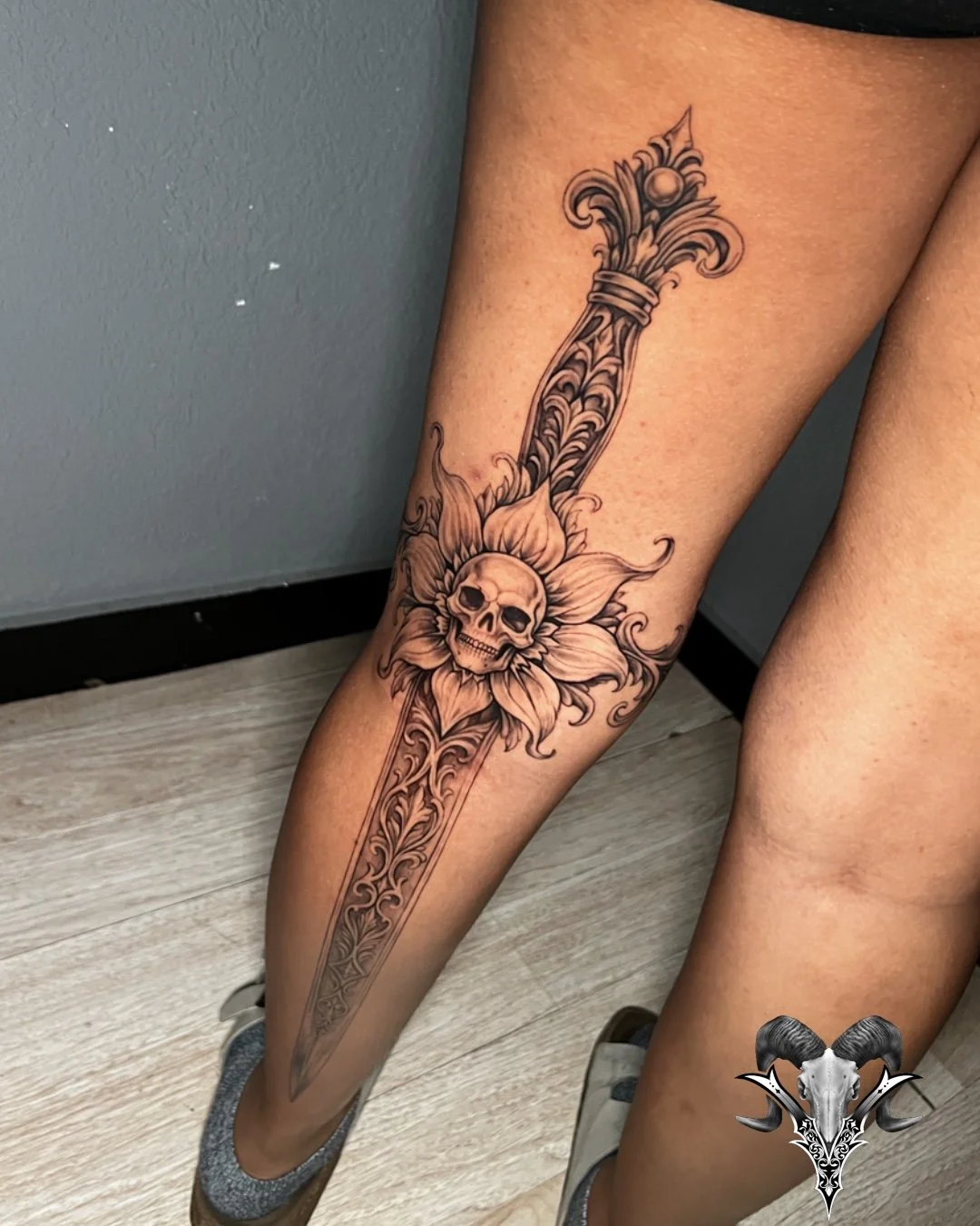 Black and grey ornamental blade tattoo with a skull and flower on the leg by Mi Vida Tattoos