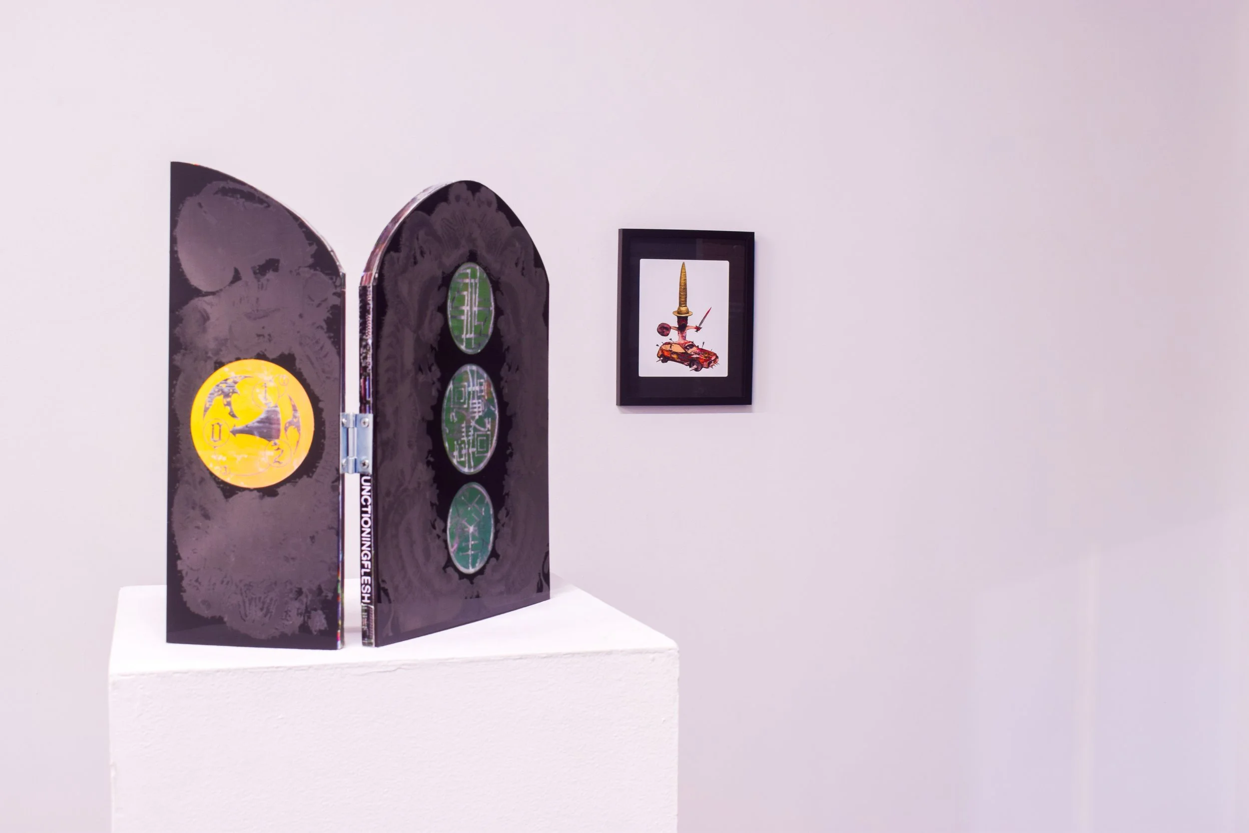 An art installation features a black folding screen with three green circular patterns and a yellow sticker, placed on a white pedestal, with a framed artwork of a sword stabbing a car on the white wall behind.