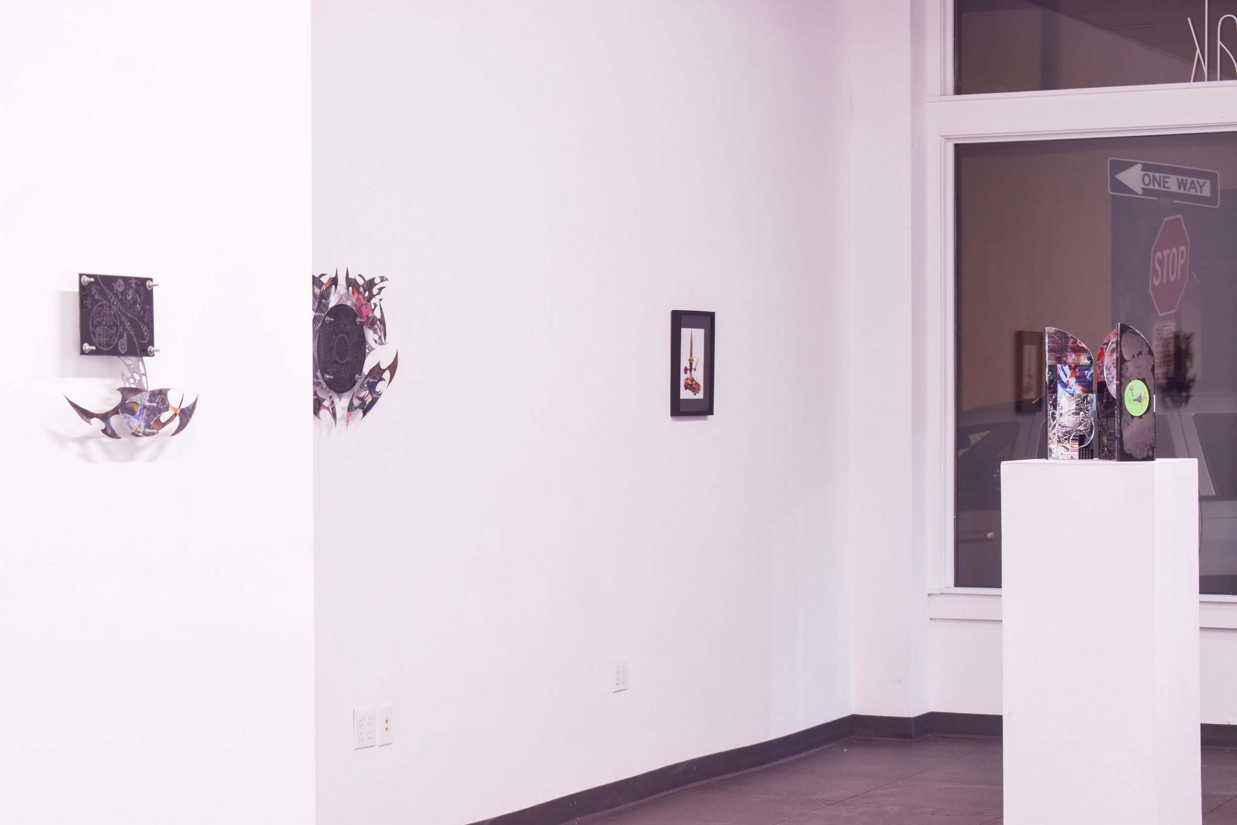 Art gallery with wall sculptures and framed artwork, and a pedestal displaying colorful abstract sculptures near a large window.