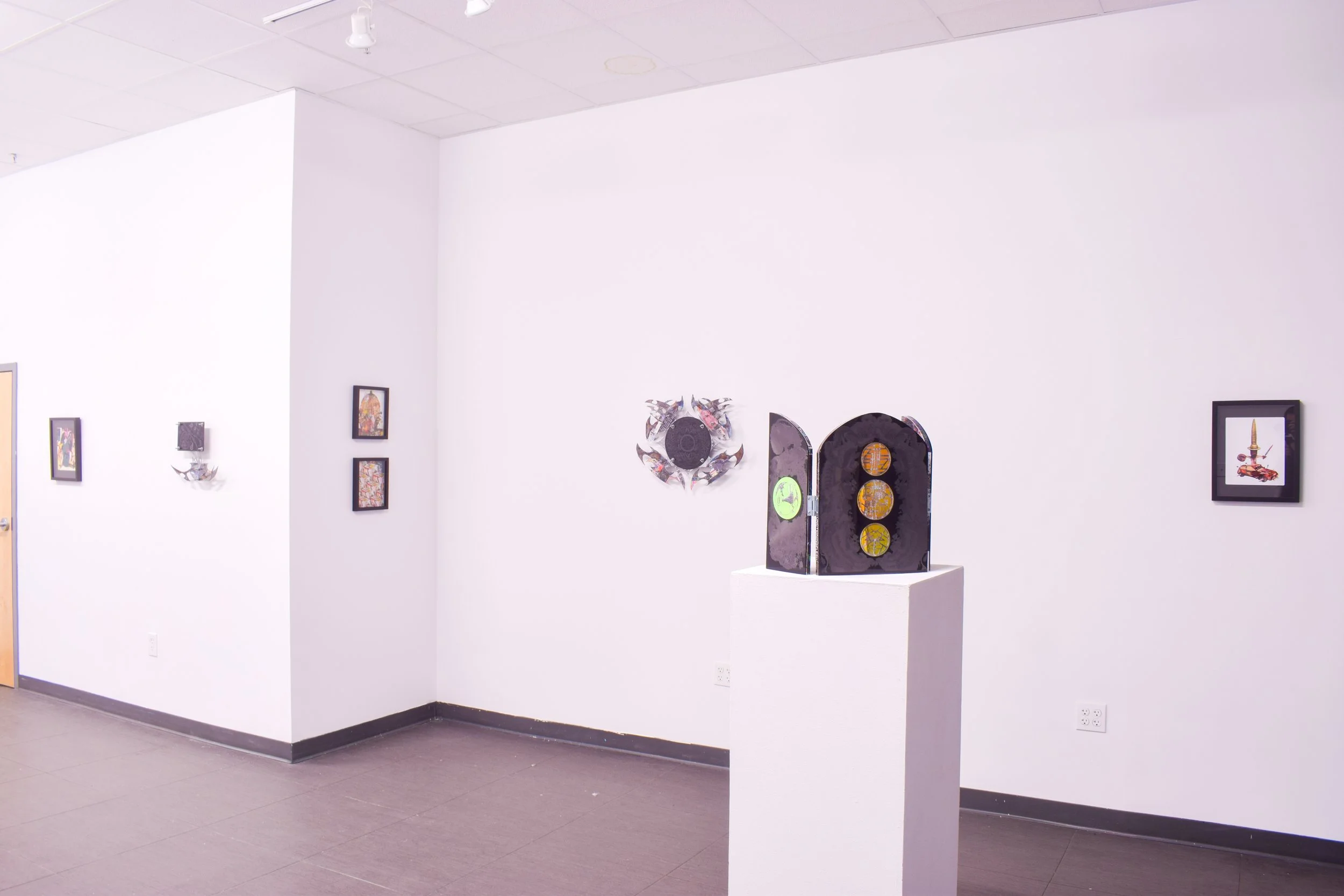 Art gallery with framed artworks on white walls, a sculpture on a pedestal, and a circular wall decoration.