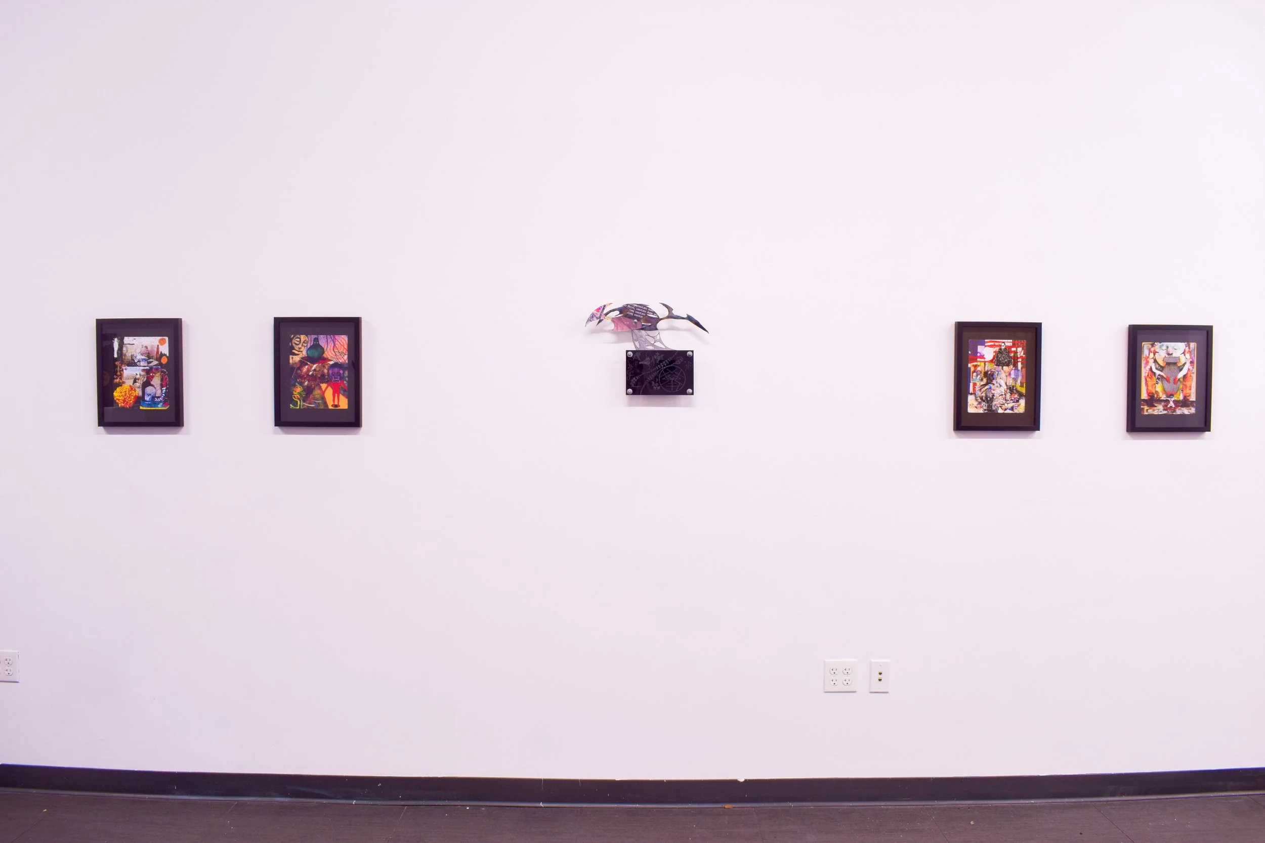 Art gallery with five framed colorful artworks on a white wall and a metal sculpture of a fish above the central piece.