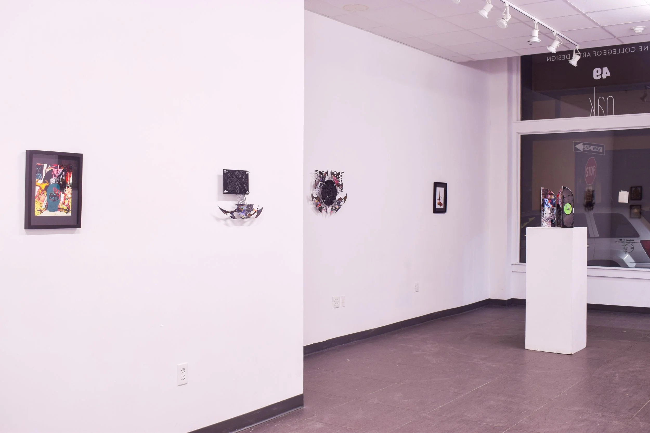 Art gallery with several small framed artworks on a white wall, some sculptures on a white pedestal near a large window showing street signs, parked car, and other artworks.