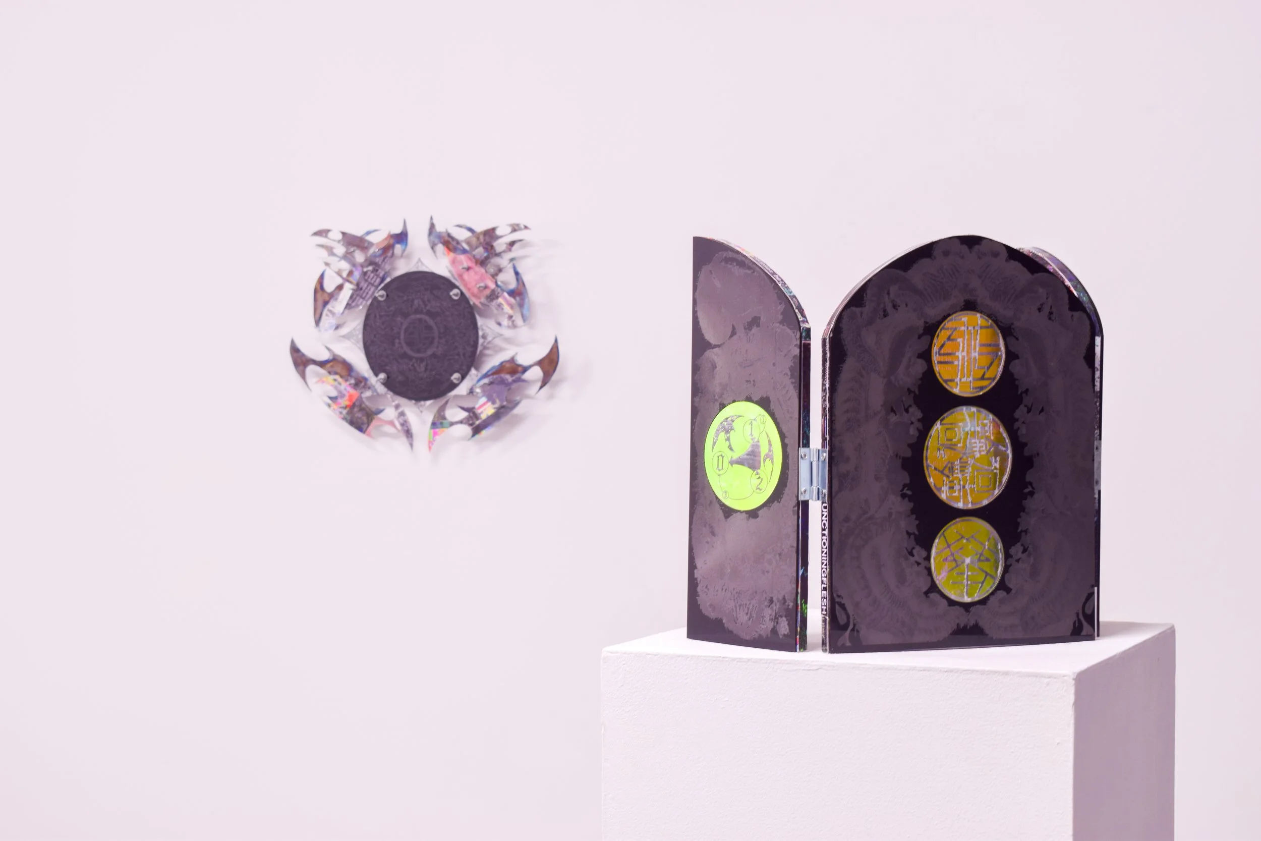 Two black and gold decorative panels with circular illustrations stand on a white pedestal, with a circular geometric-shaped art piece on the wall to the left, against a plain light background.