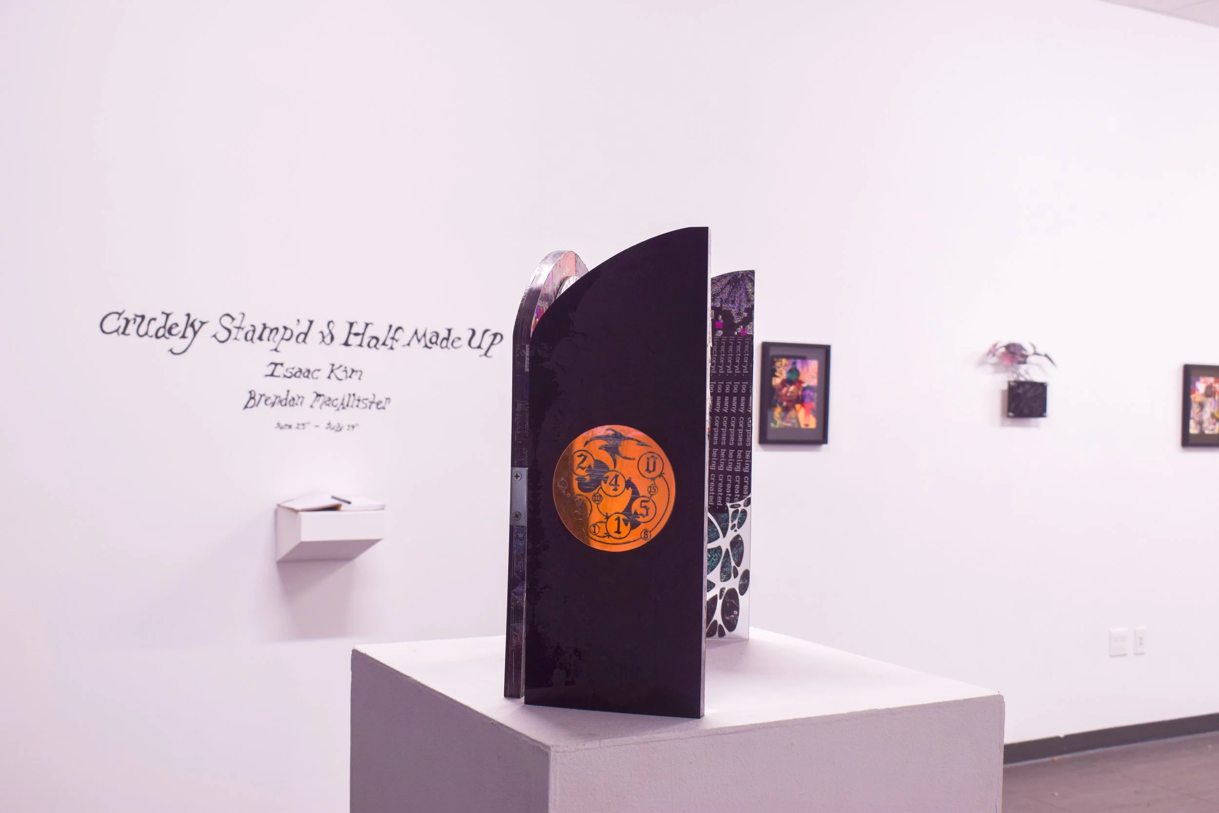 Art exhibit with a black sculpture featuring an orange circular design, displayed on a white pedestal in a gallery with framed artworks on white walls.