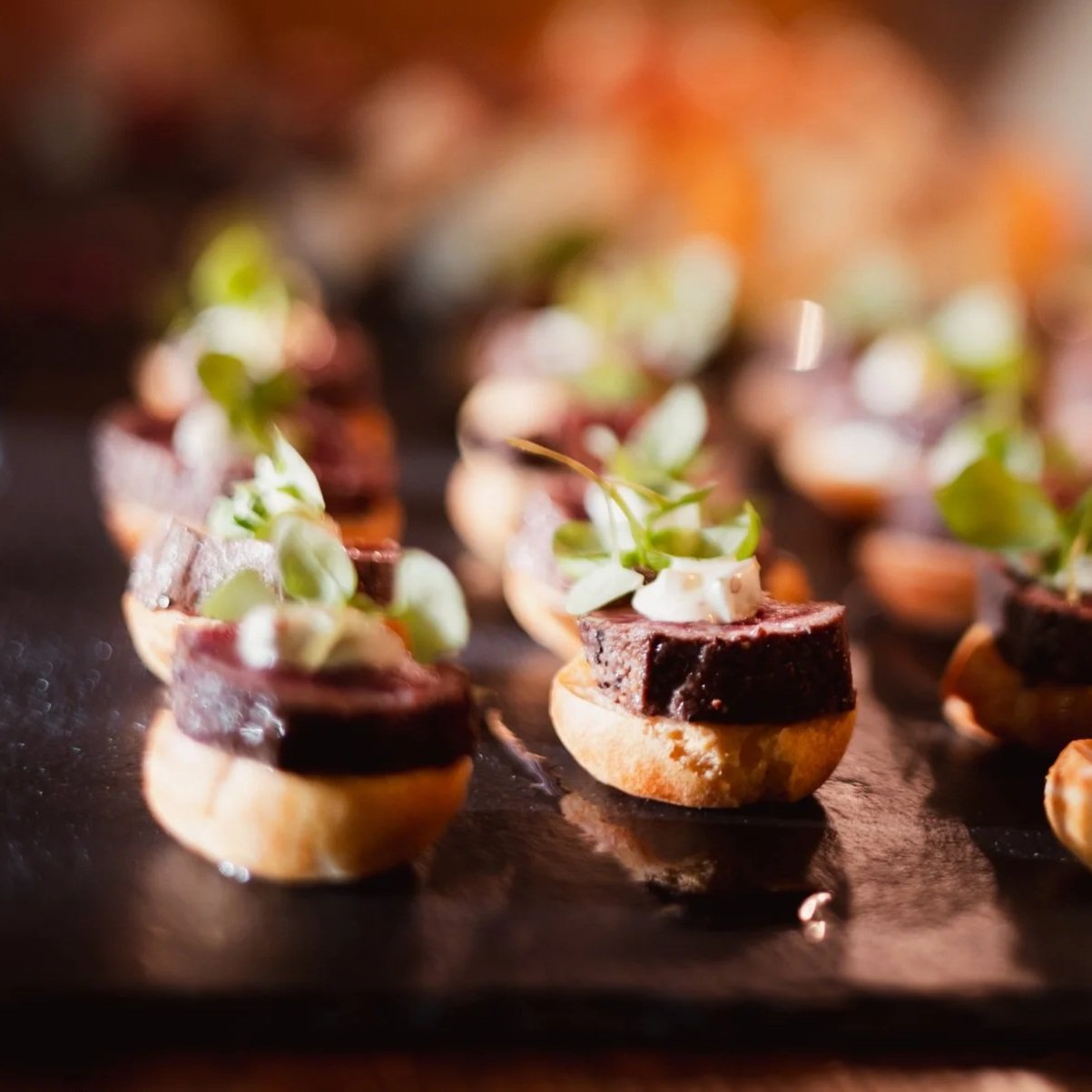 A server holding a tray of small appetizers in tart shells, topped with orange-colored food, possibly smoked salmon and garnishes, at a dimly lit event.