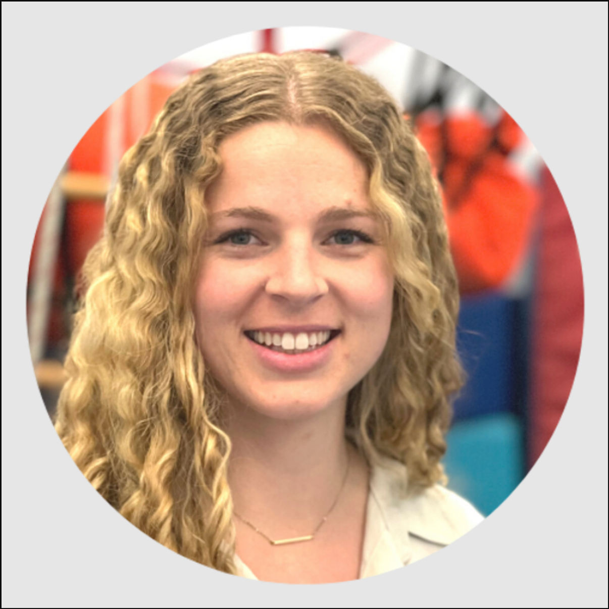 A woman with curly blonde hair smiling, wearing a light-colored top and a delicate gold necklace, with a blurred indoor background.