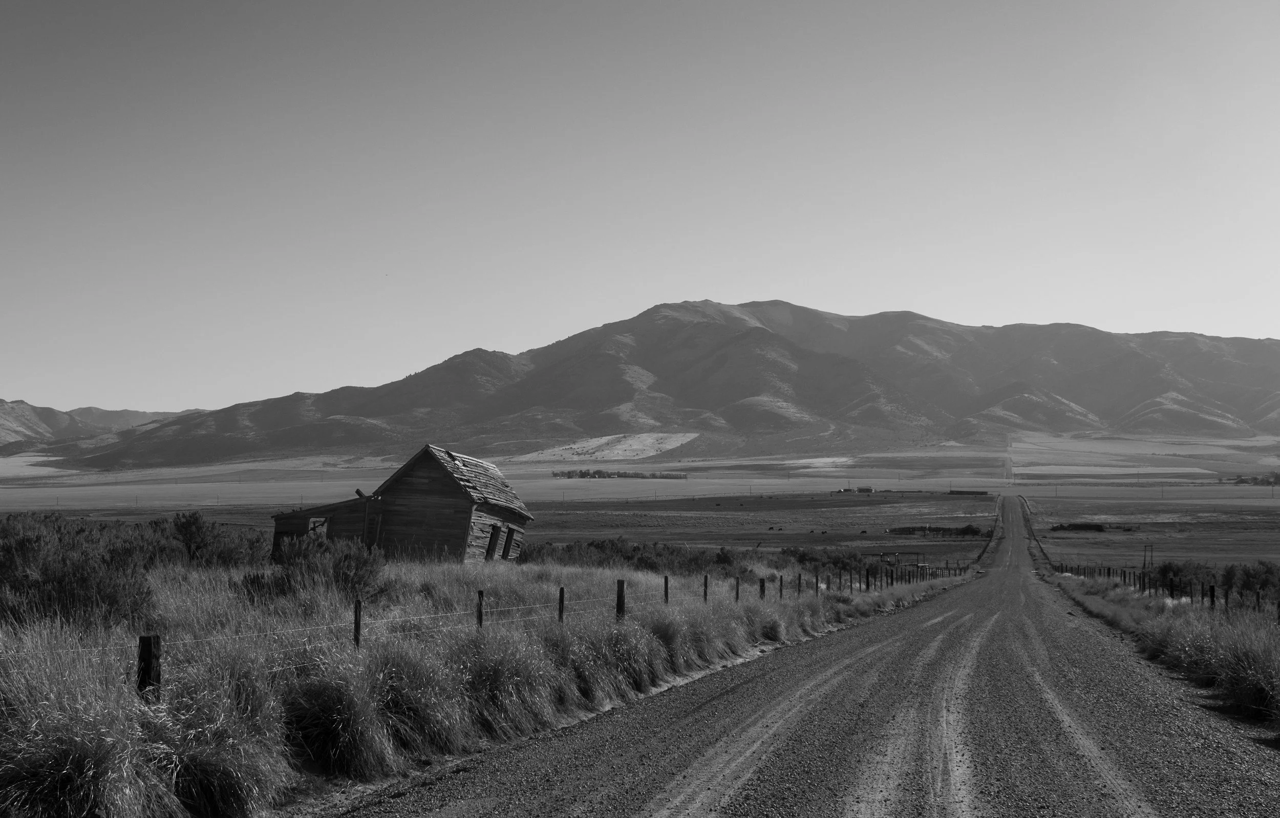 Black and white photo of a rural landscape with a dirt road, an old leaning barn, fence posts, grassy fields, and distant mountains.