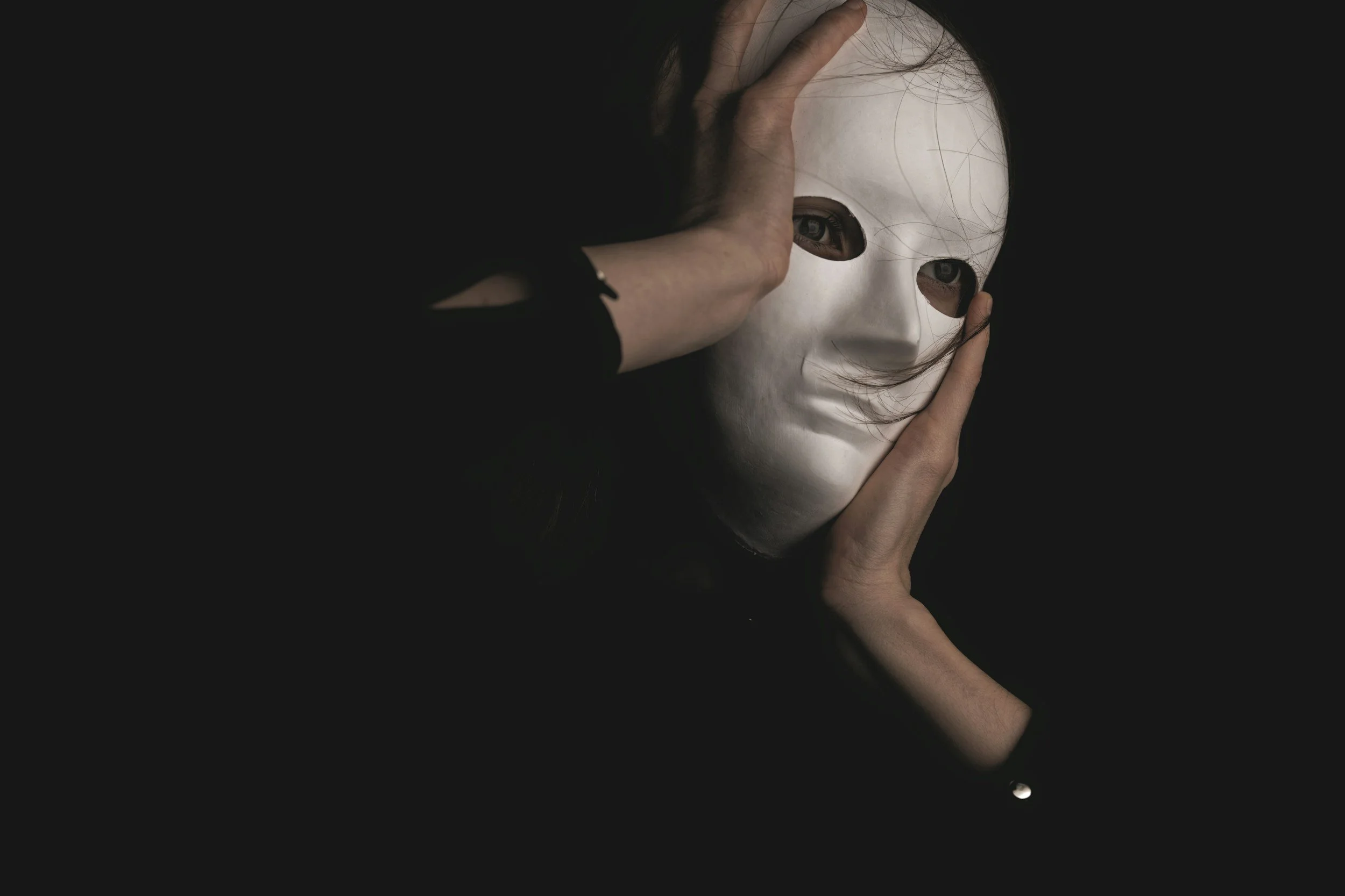 A person holding a white mask with dark eye holes, while the mask is being held up against the person's face, with dark hair falling around the mask.