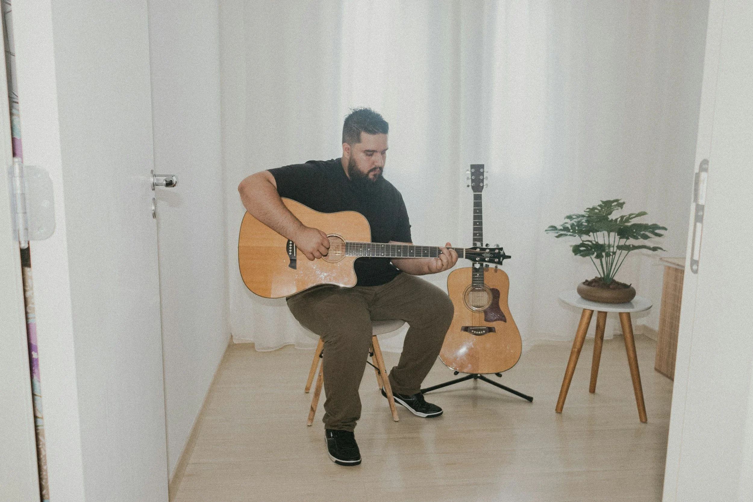 A man with a beard sitting on a chair in a white room, playing an acoustic guitar. There are two other guitars behind him and a potted plant on a small table to his right.