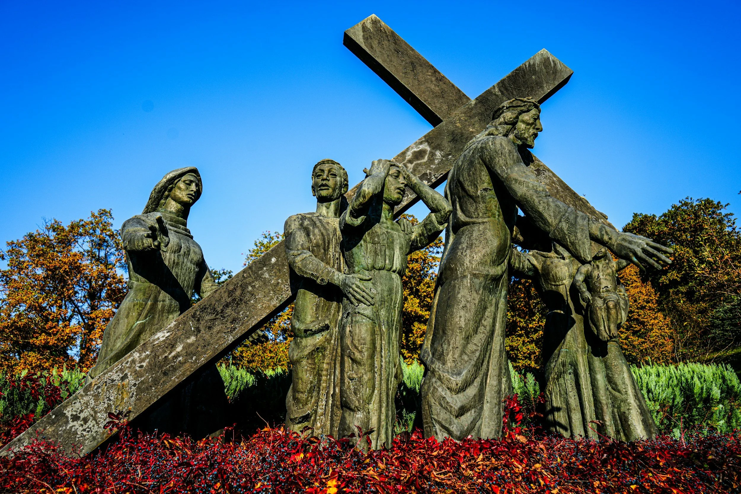 Statue of Jesus Christ carrying a cross, with two women figures on either side, set against a blue sky with trees and fallen leaves.