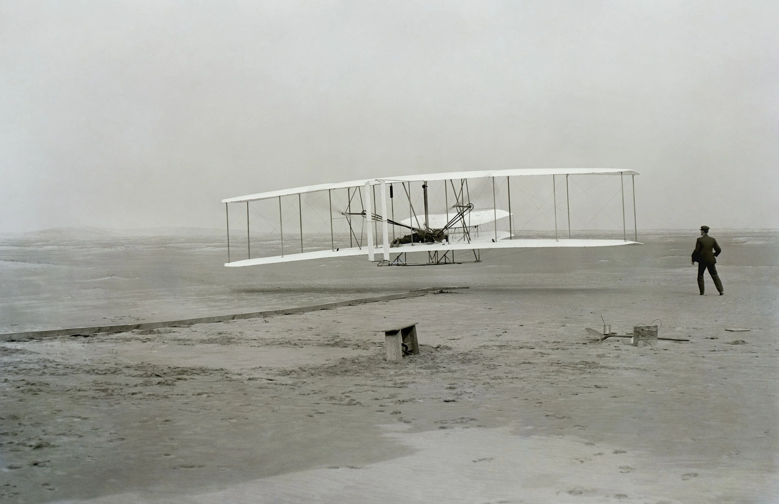 A vintage early 20th-century airplane with large wings and exposed framework on a flat, barren landscape under a cloudy sky, with a person standing nearby.