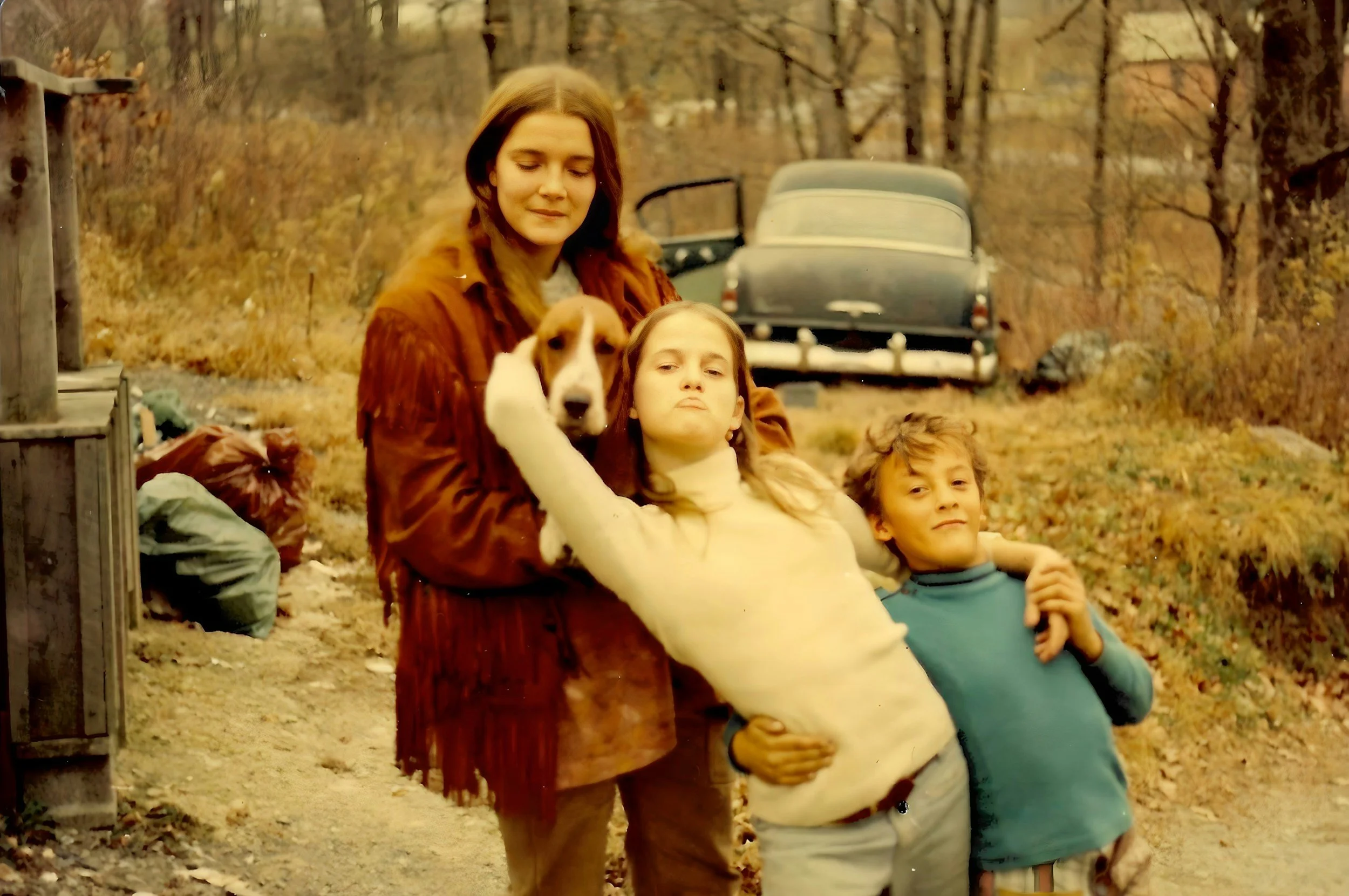 A woman with long brown hair wearing a red jacket holds a puppy with white and brown fur. Two children, a girl with blonde hair and a boy with brown hair, stand beside them outdoors during autumn, in front of leafless trees and a vintage car.