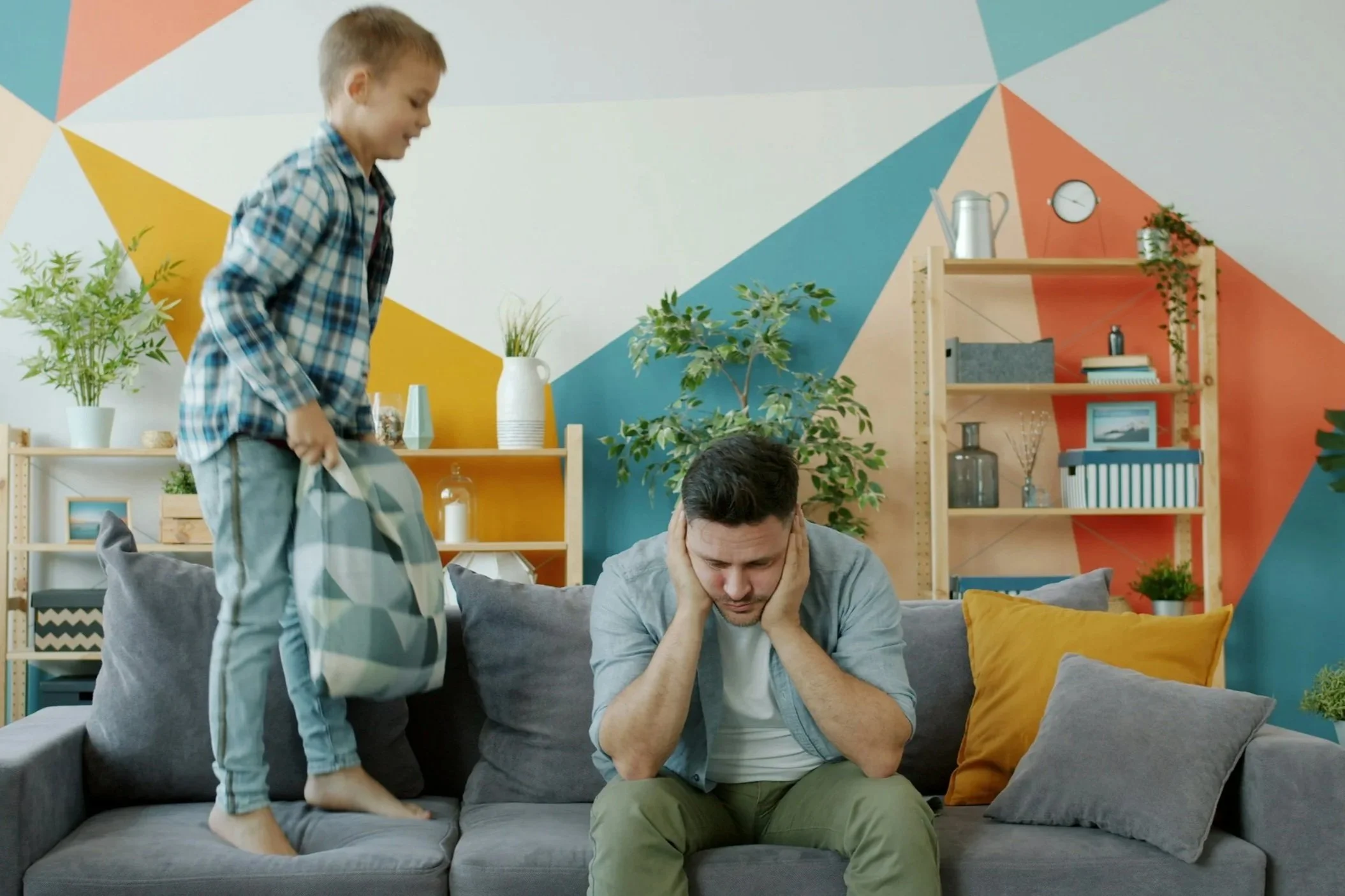 A boy standing on a sofa looking at a man sitting with his head in his hands, appearing stressed or upset, in a colorful living room with plants and artwork.