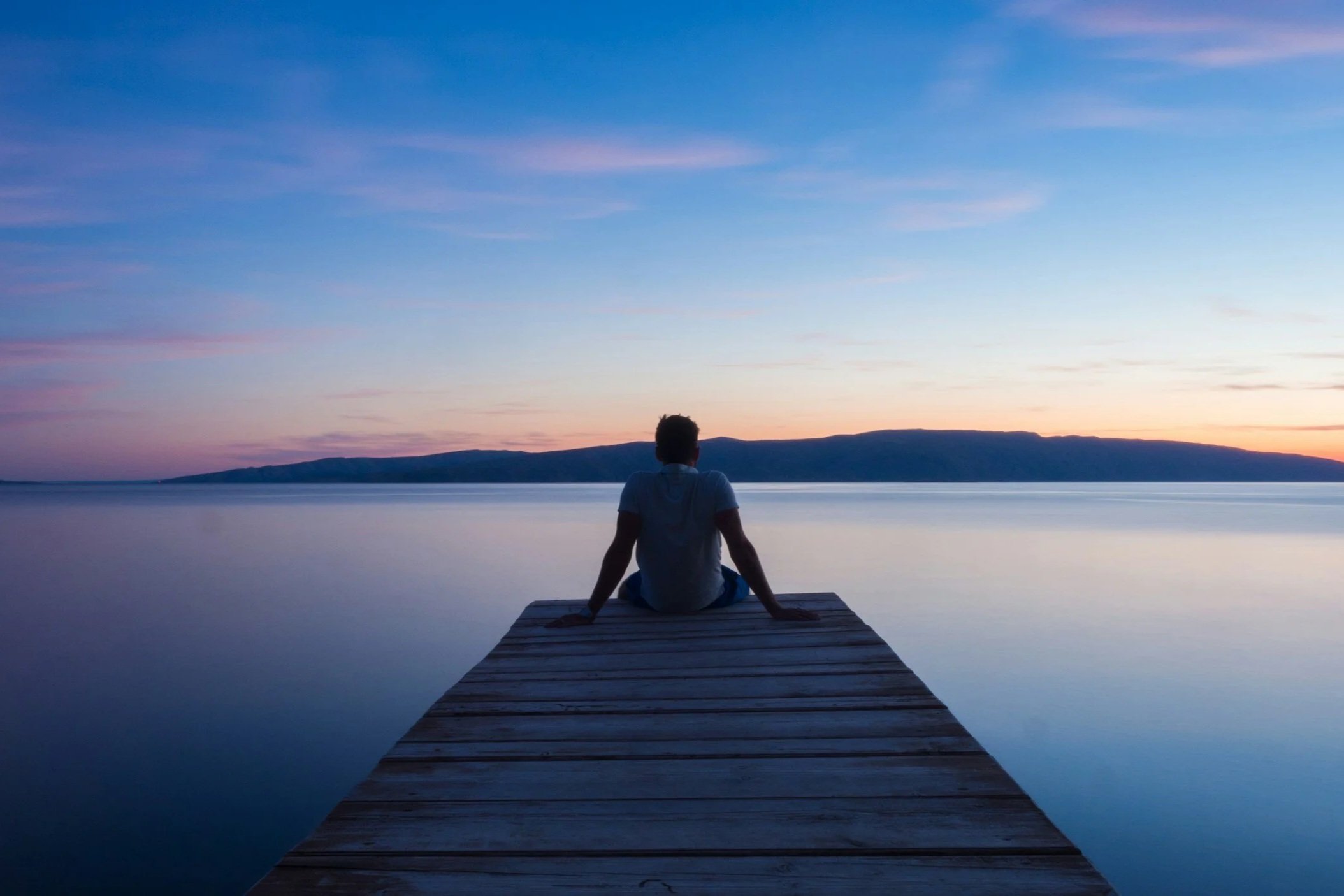 Person sitting on a wooden dock overlooking calm water during sunset with distant mountains and a colorful sky.