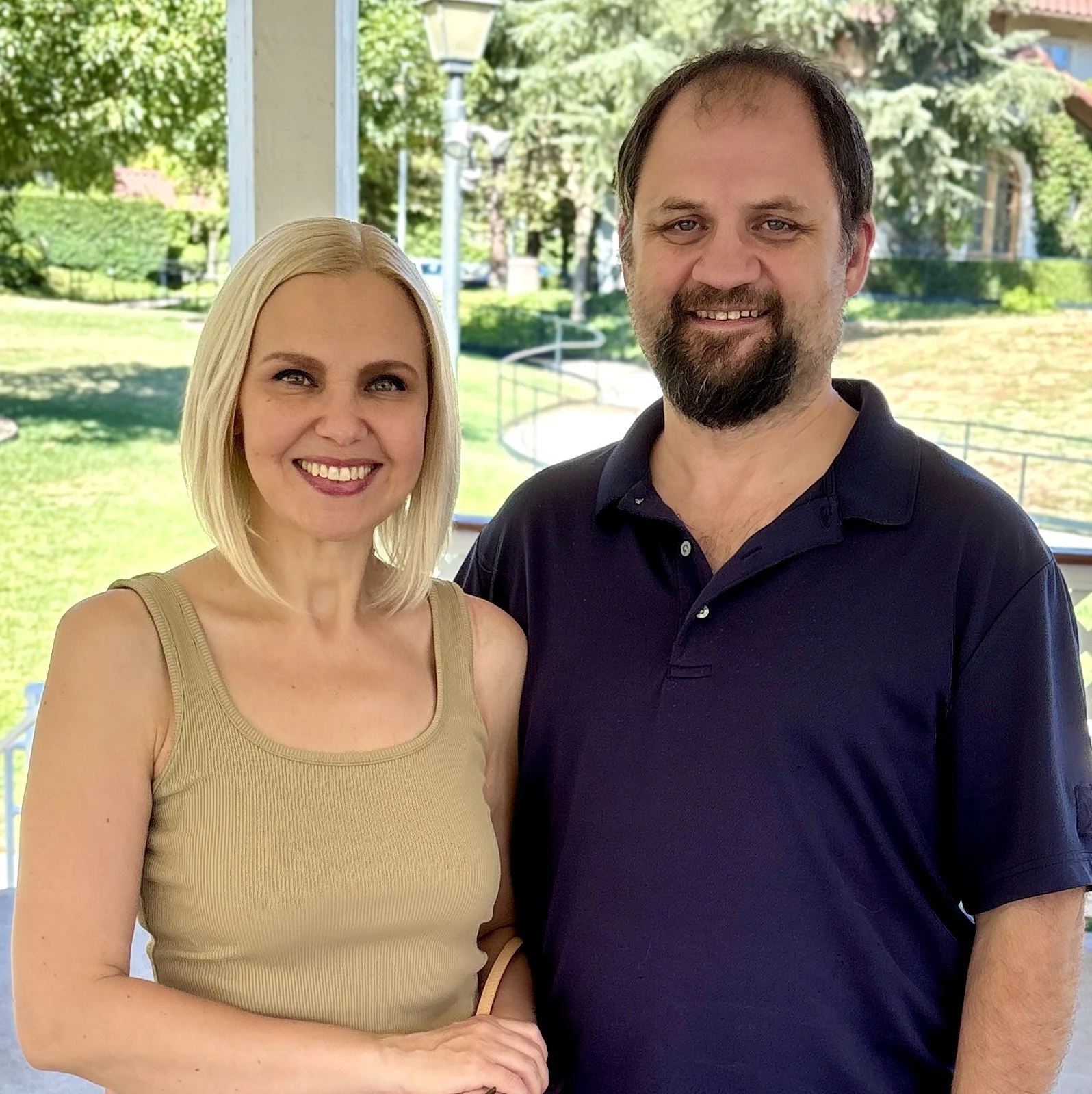 A woman with blonde hair wearing a beige tank top and a man with brown hair and beard in a dark polo shirt, standing outdoors on a porch with greenery in the background.
