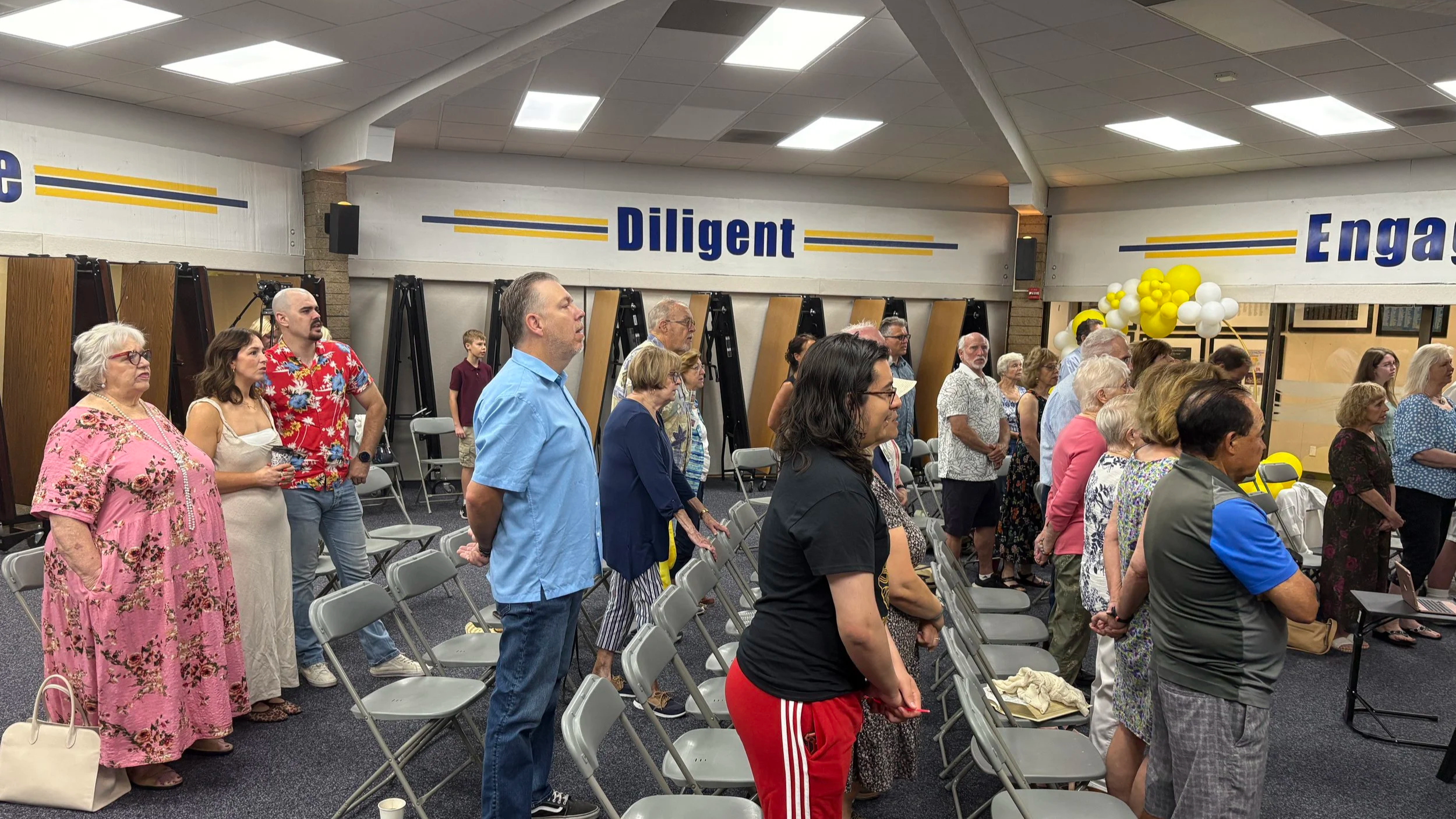 People attending an indoor event, standing and facing forward, with some chairs arranged in rows. The room has signs on the walls reading 'Diligent' and 'Engaged,' and decorations including balloons in yellow and white.