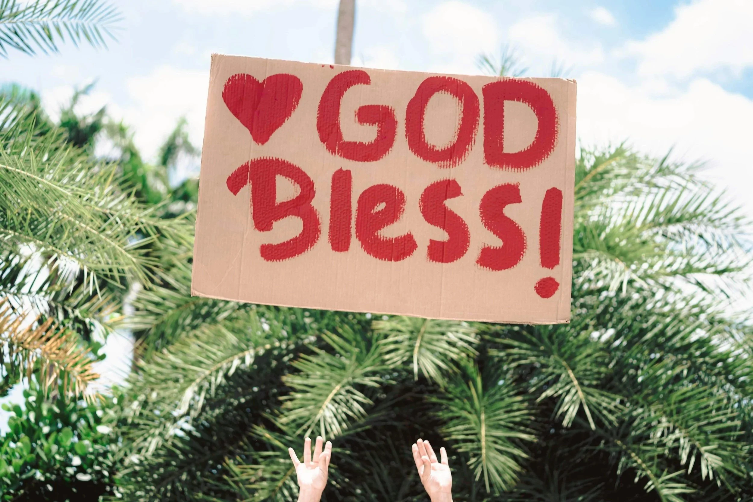 People holding a cardboard sign that says 'Good Bress!' with a heart symbol, raising their hands against a backdrop of trees and partly cloudy sky.