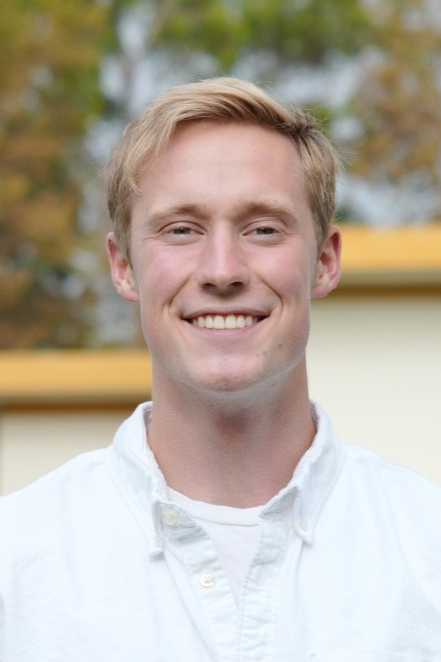 A young man with blonde hair smiling outdoors, wearing a white button-down shirt, with trees and a building in the background.