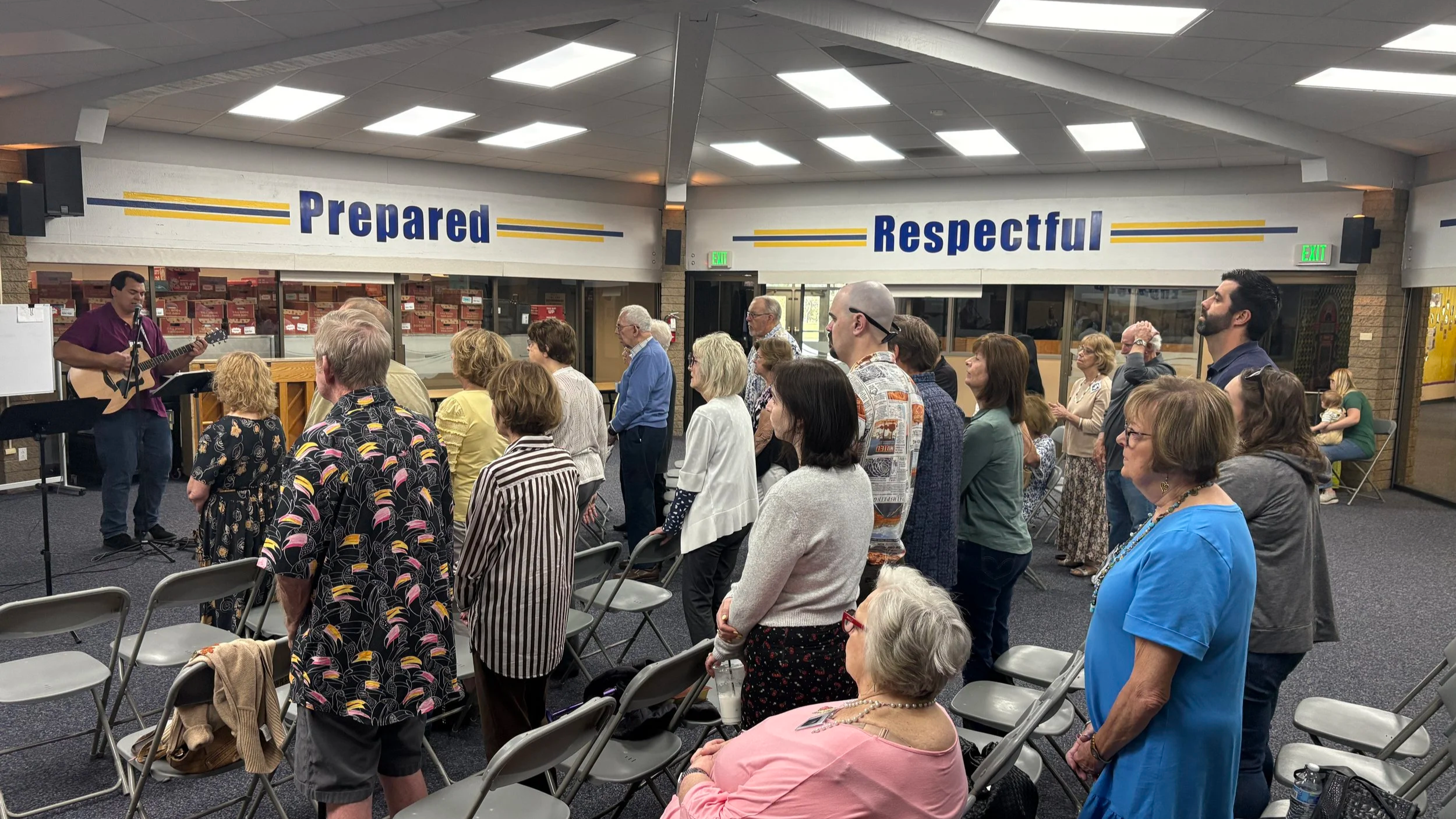 A group of people standing and participating in a gathering, some are singing or listening to a man playing guitar and singing. The background has two large signs saying 'Prepared' and 'Respectful'.