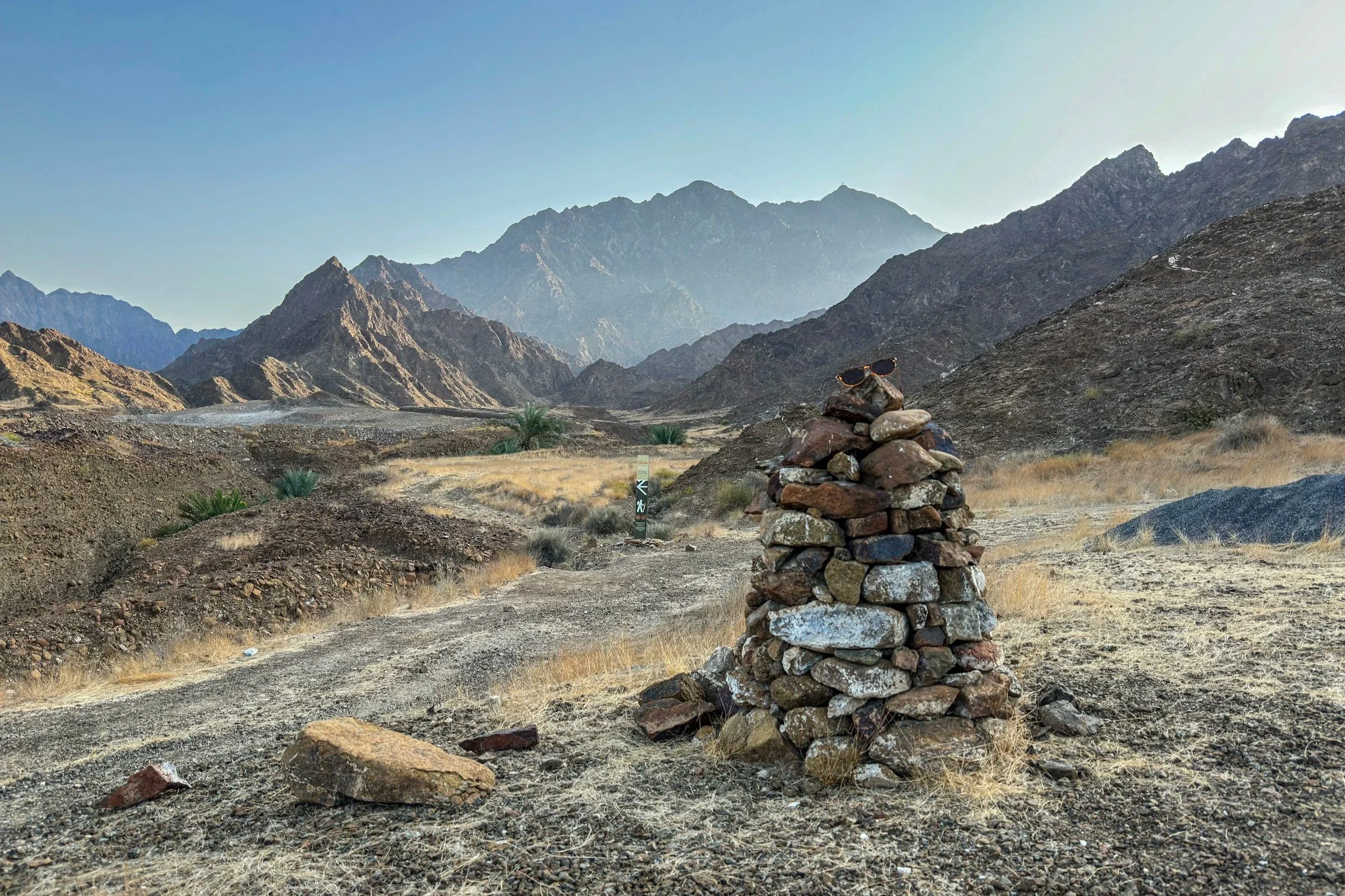 A dry mountainous desert landscape with rugged peaks in the background, a dirt path in the foreground, some sparse vegetation, and a cairn with sunglasses resting on top.