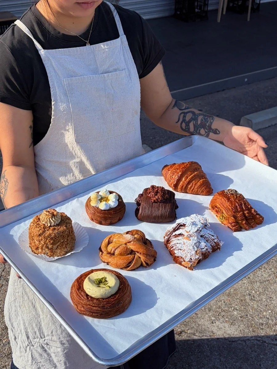 A baker holding a tray of Time & Temp pastries including a croissant, cardamom bun, passionfruit tart, and other baked goods on a white paper-lined tray in the morning sun.