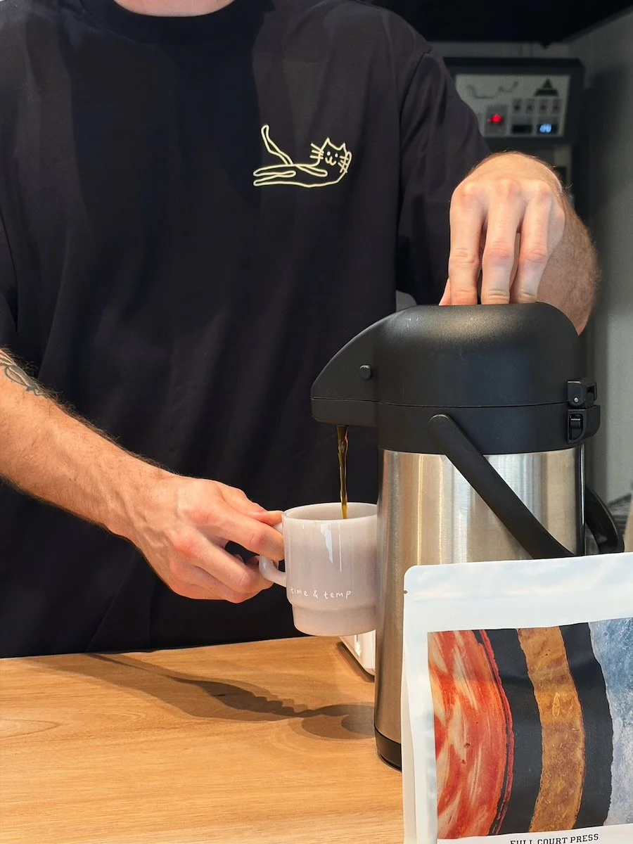 A person in a navy shirt with Time & Temp cat drawing logo is pouring coffee into a mug from a thermal coffee dispenser.