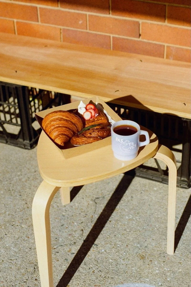 A wooden stool with a tray holding a croissant, a strawberry danish, and a pink Time & Temp coffee mug of coffee in front of a brick wall.
