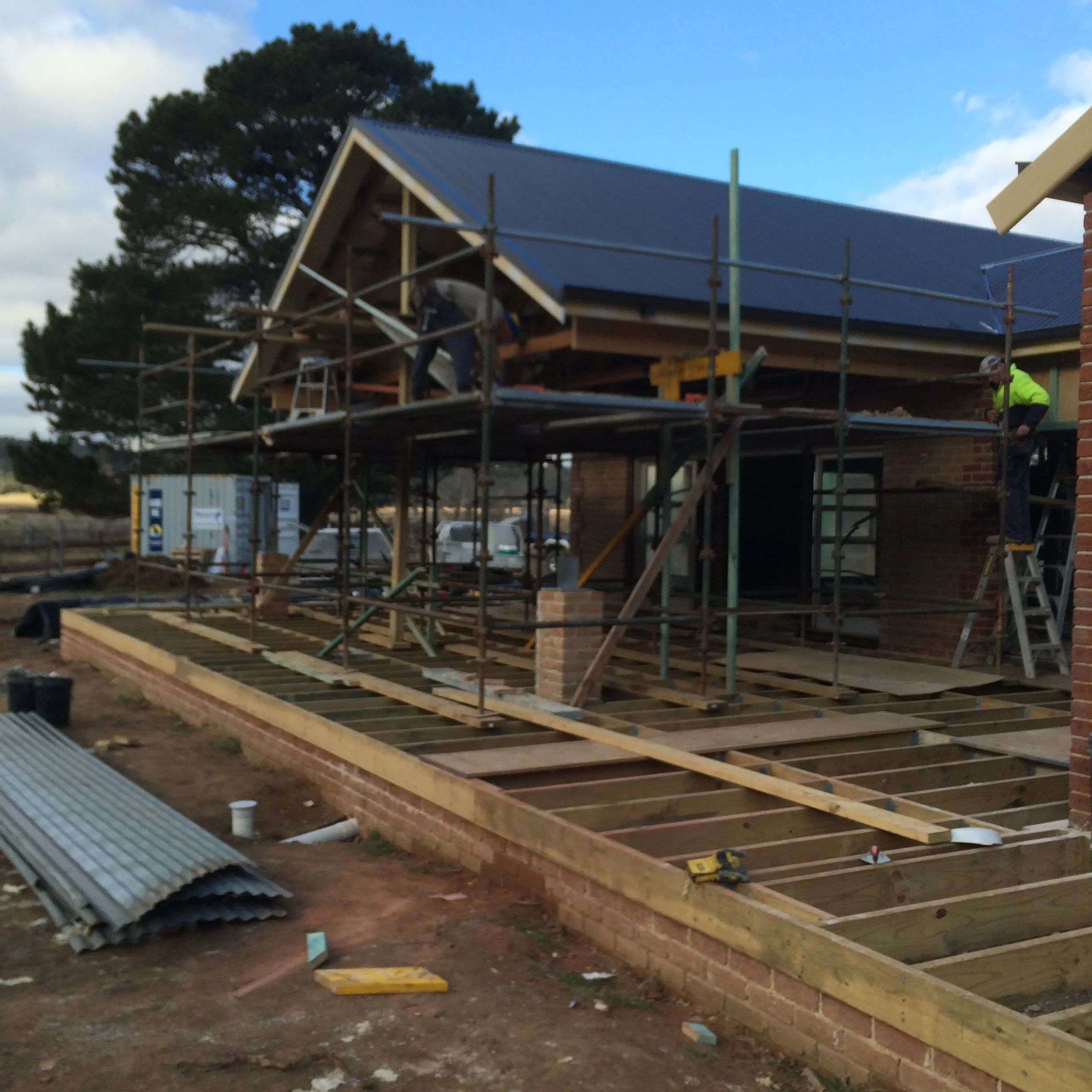 House under construction with scaffolding and workers, surrounded by trees and a clear sky.