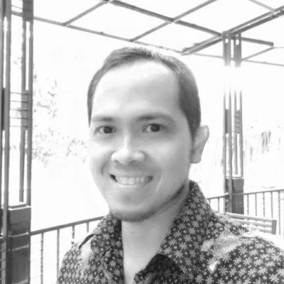 A smiling man with short dark hair and a patterned shirt, standing outdoors on a balcony or terrace with a metal railing and overhead structure.