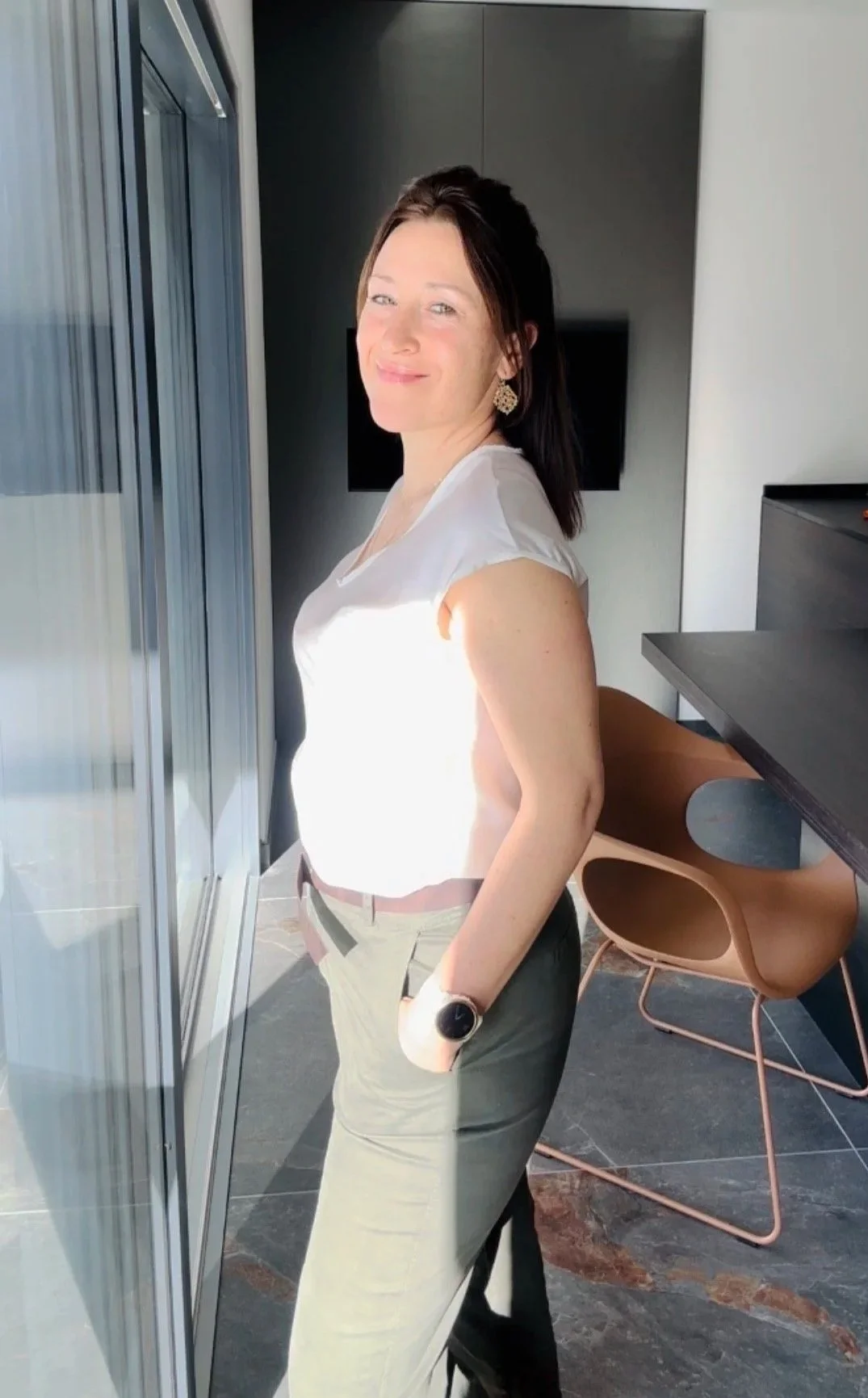 Eine Frau steht neben einem Fenster in einem modernen Raum, lächelt und trägt eine weiße Bluse, eine beige Hose, eine Uhr und große, goldene Ohrringe.
