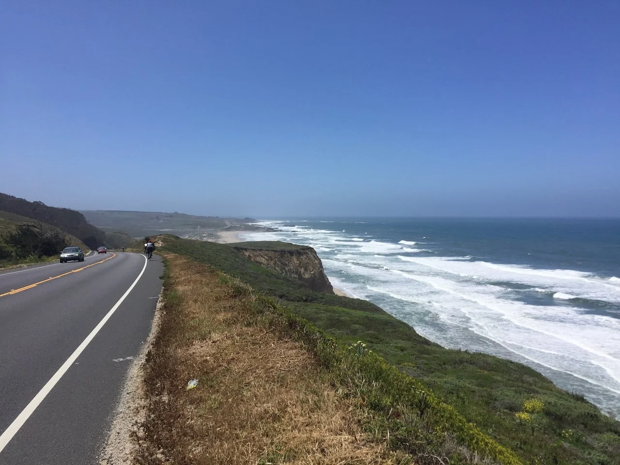 A winding coastal road alongside a green cliff overlooking the ocean with waves and a clear blue sky.