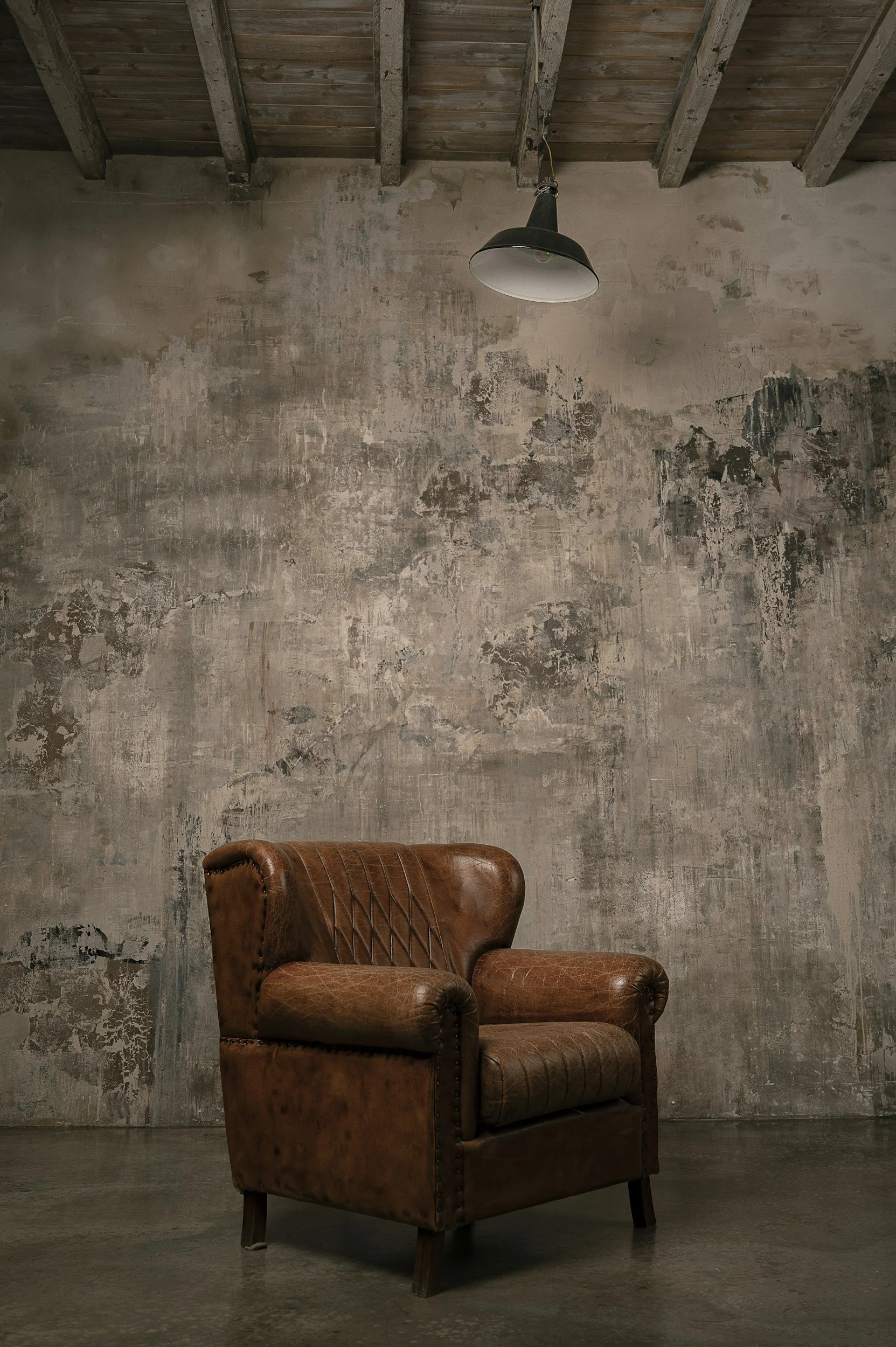 A brown leather armchair with a diamond quilted design on the backrest, sitting in front of a worn, textured gray wall and a rustic ceiling with exposed wooden beams and a single black hanging lamp.