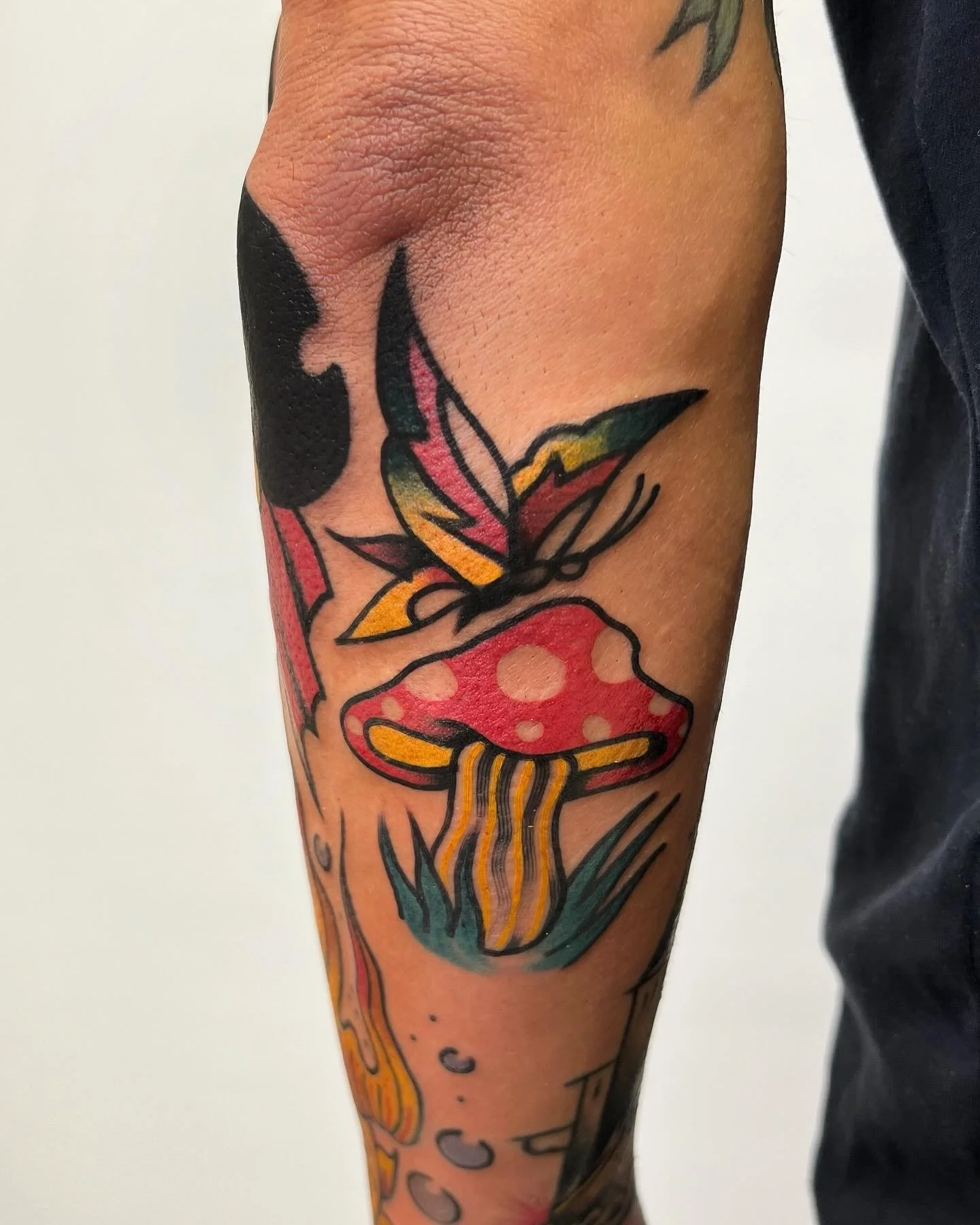 Tattoo of a red mushroom with yellow gills and a pink-spotted cap, alongside a colorful butterfly with pink, yellow, and dark blue wings, on a person's arm.