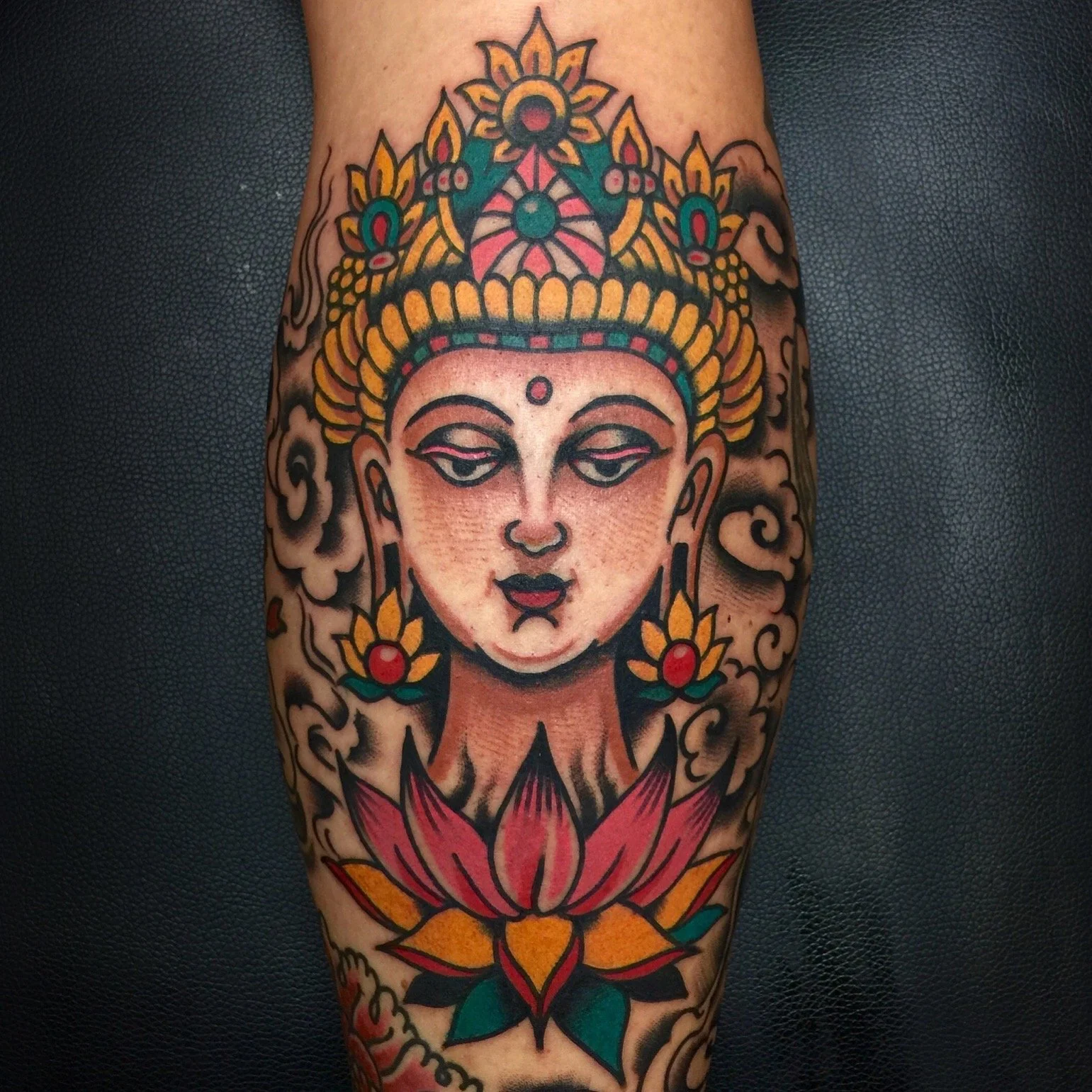 Tattoo of a meditative female figure with a crown, surrounded by clouds, a lotus flower at the bottom, and colorful decorative elements.