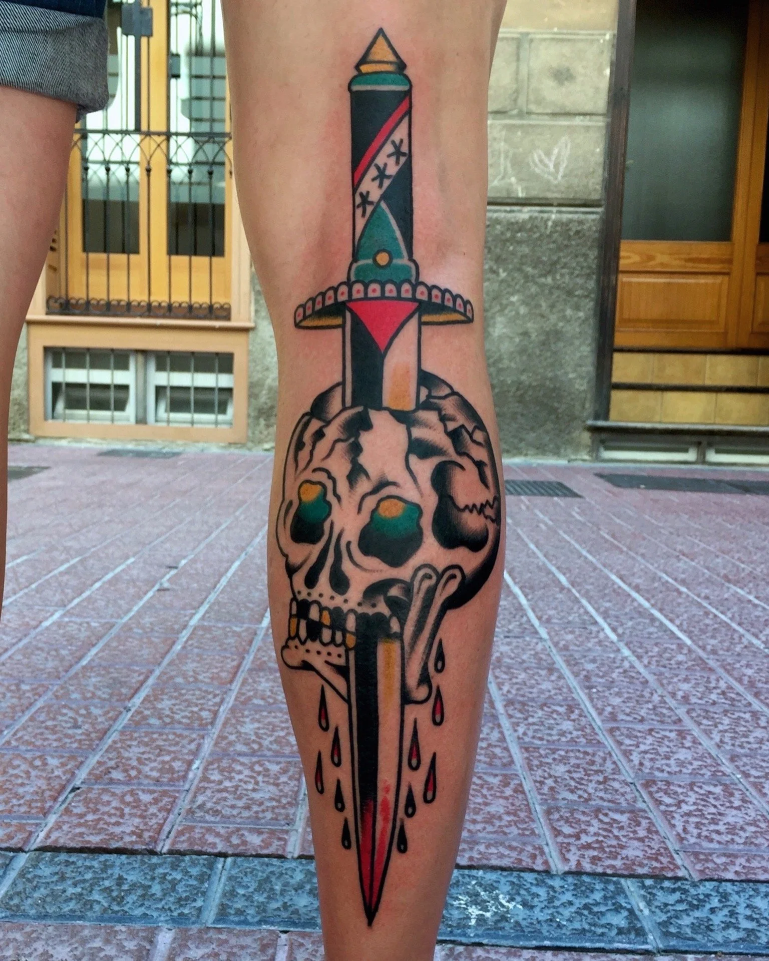 Tattoo of a skull with dark eye sockets and a sword piercing through it, the sword is decorated with colorful elements, and red drops are dripping from the blade.