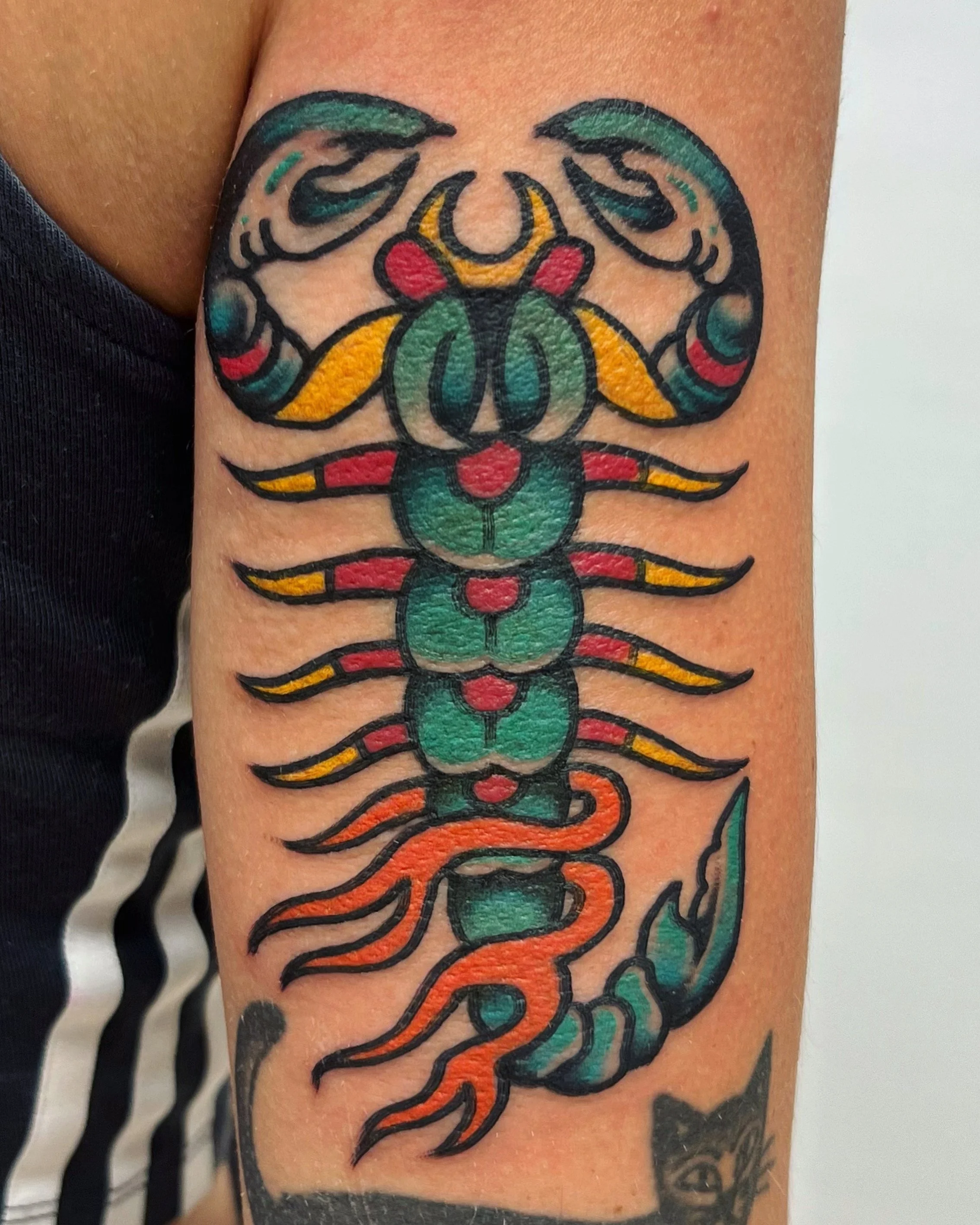 Colorful tattoo of a stylized scorpion on person's arm, featuring bold black outlines, green and orange hues, and red accents.