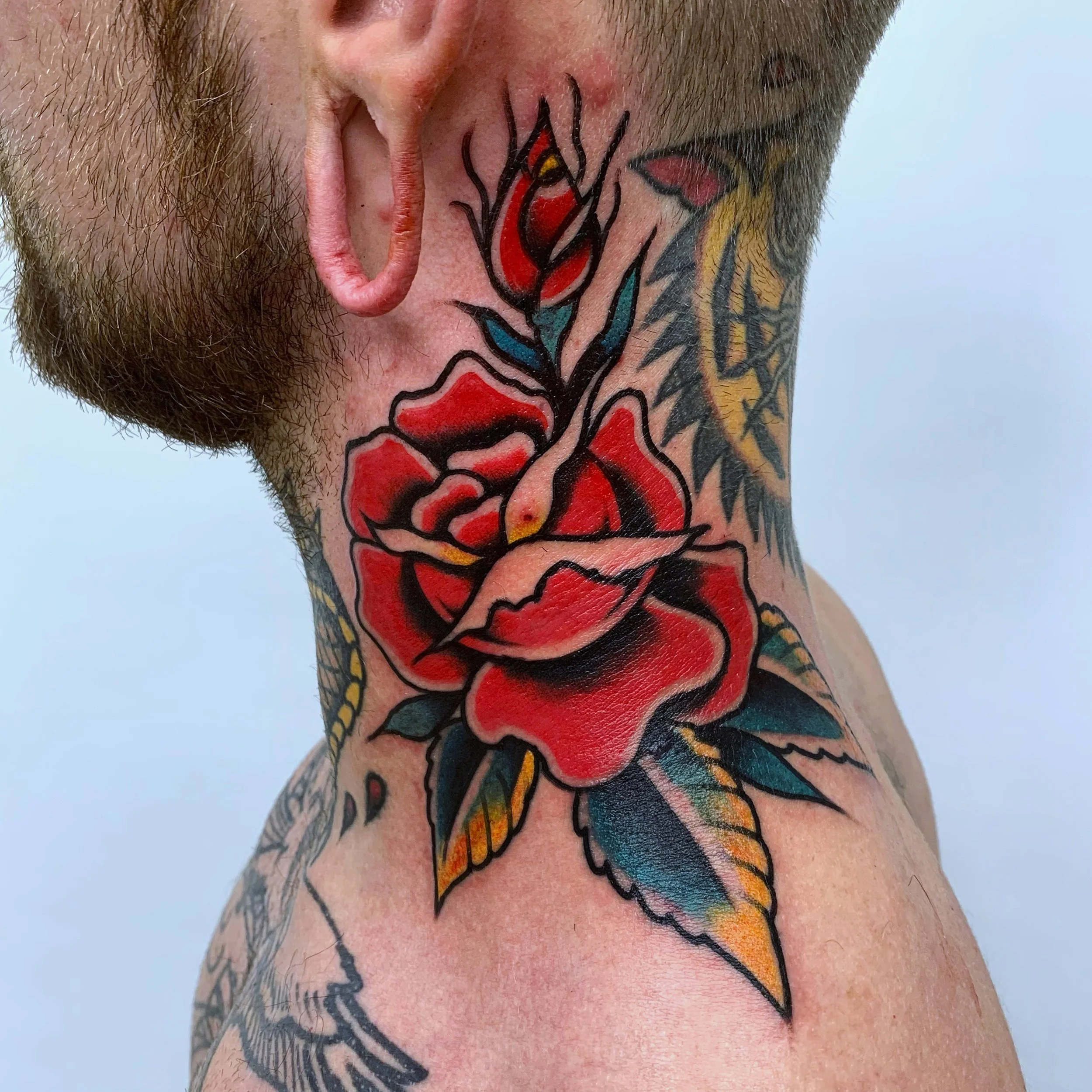 Close-up of a person's neck and shoulder showing a traditional-style red rose tattoo with green leaves. The person has a beard and a tongue piercing.