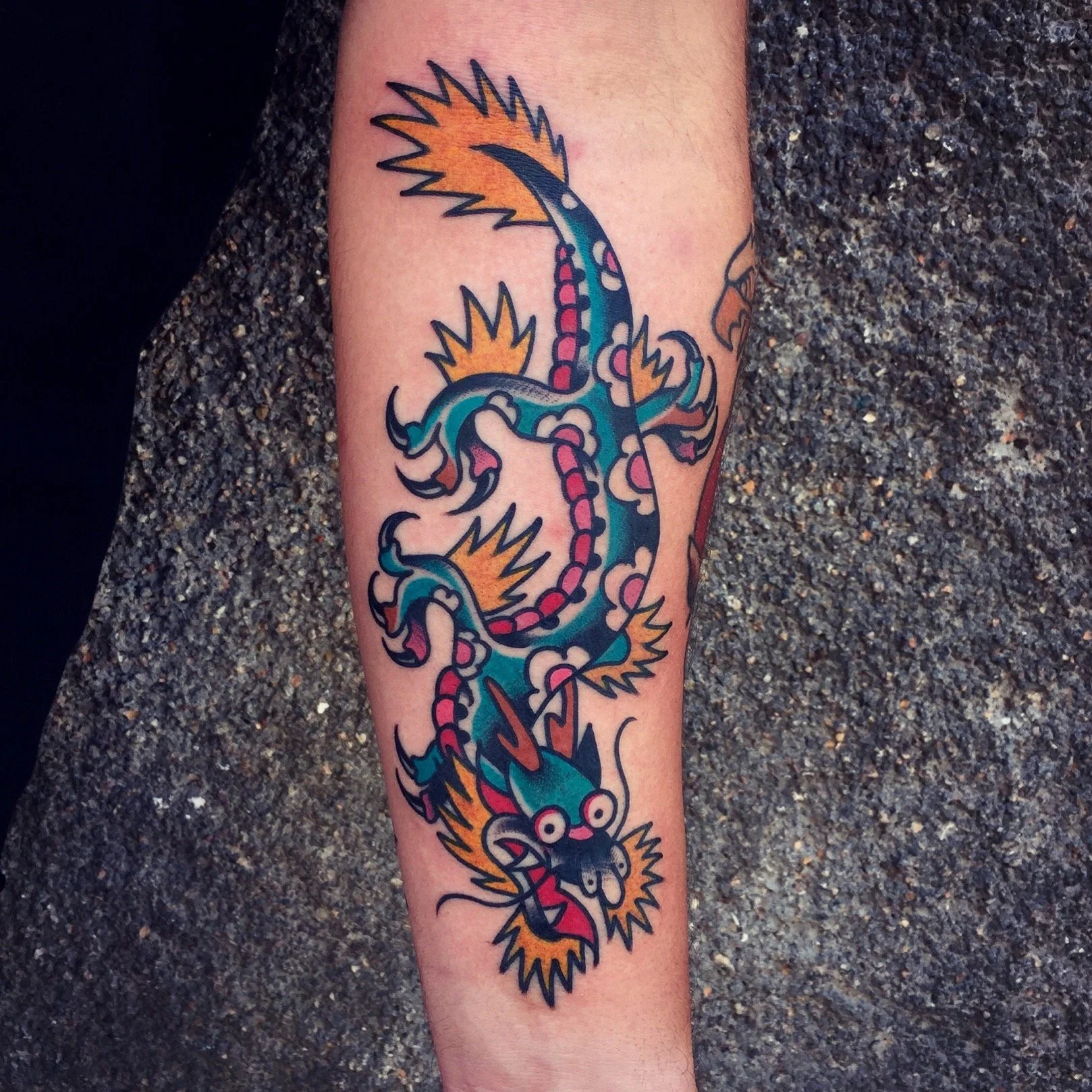 Colorful tattoo of a traditional Asian-style dragon with a long, winding body, sharp claws, and an expressive face, featuring bold black outlines and vibrant colors including blue, red, yellow, and white.