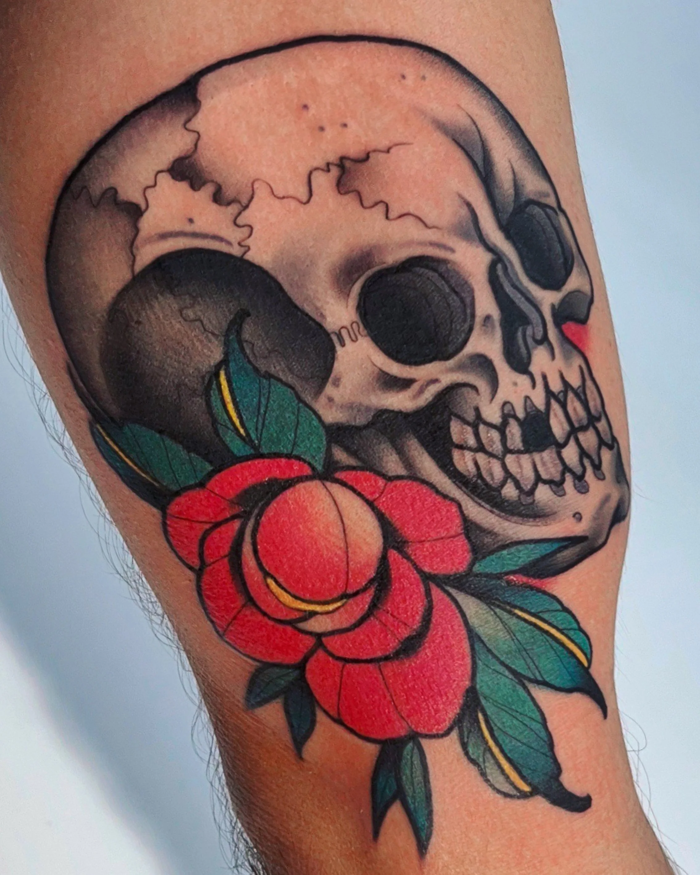 Tattoo of a skull with a red flower and green leaves on a person's arm.