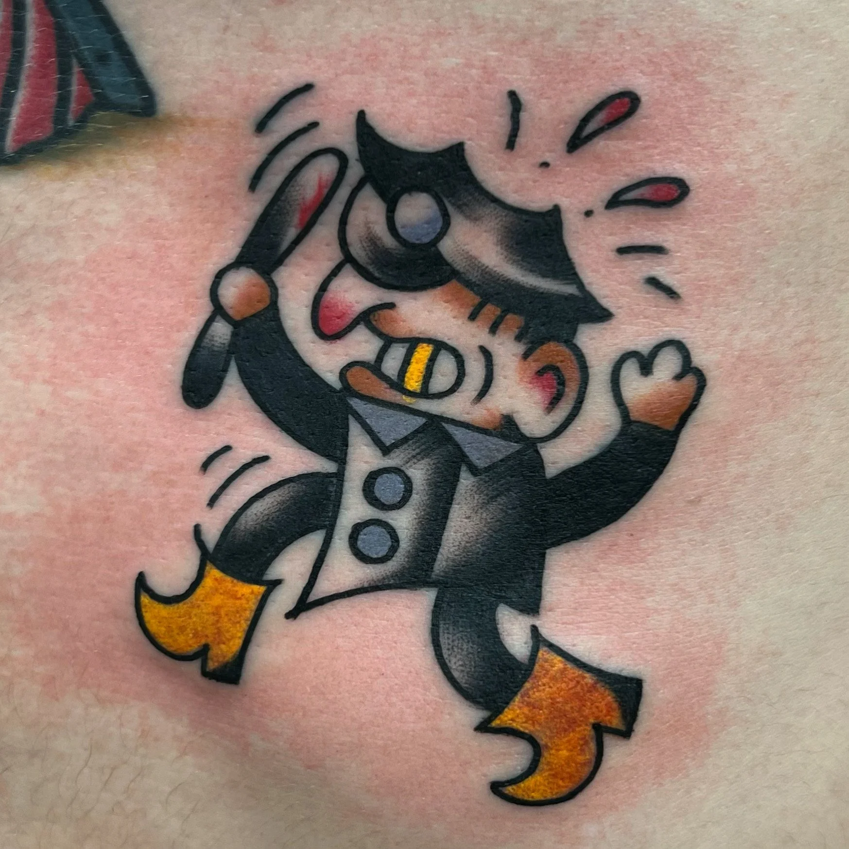 Colorful tattoo of a cartoon devil with black hair, red ears, a yellow-and-black striped tongue, and orange boots, holding a trident and making a salute gesture.